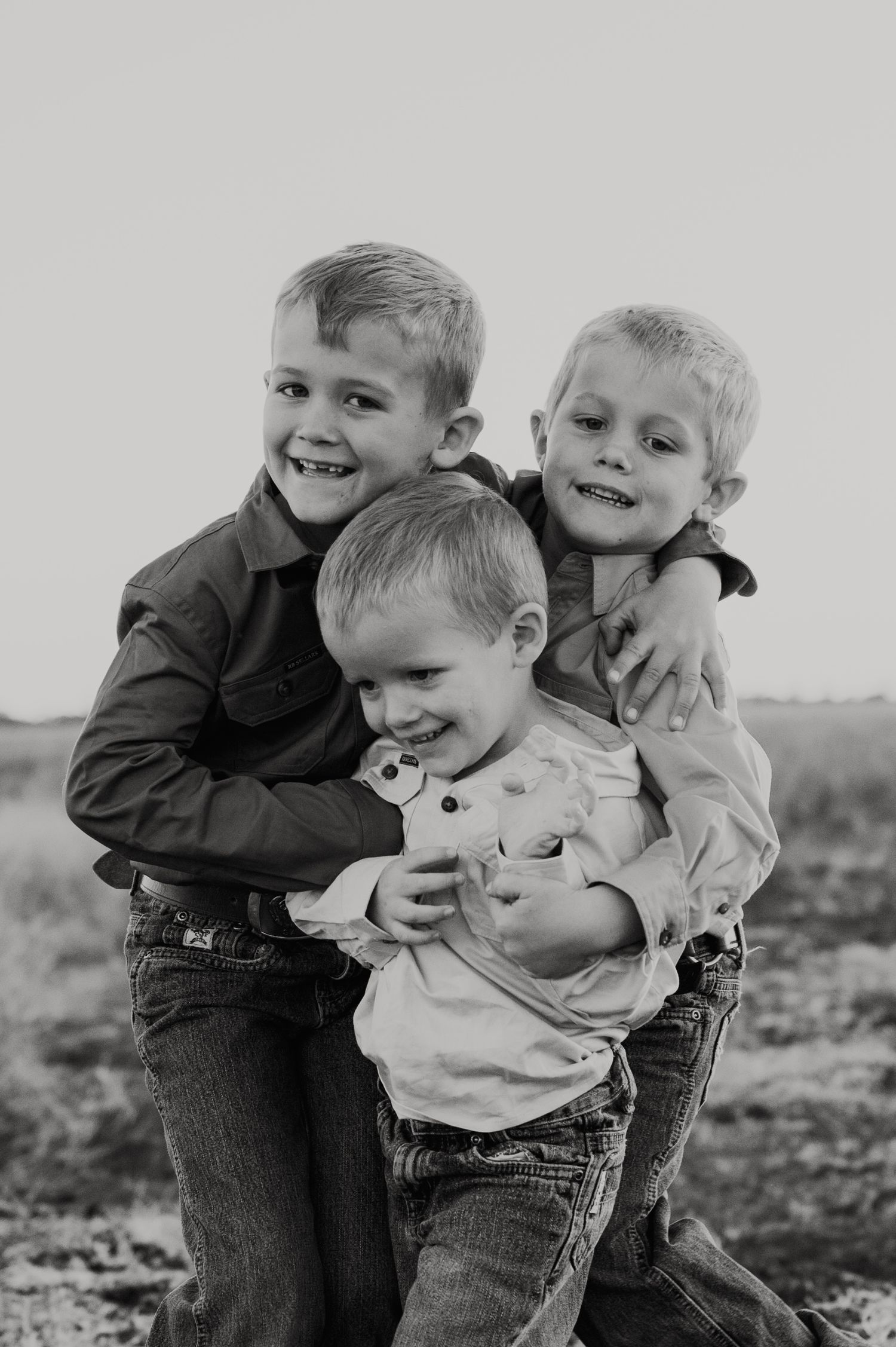 Black and white photo of three young boys hugging and smiling outdoors in a candid family portrait.