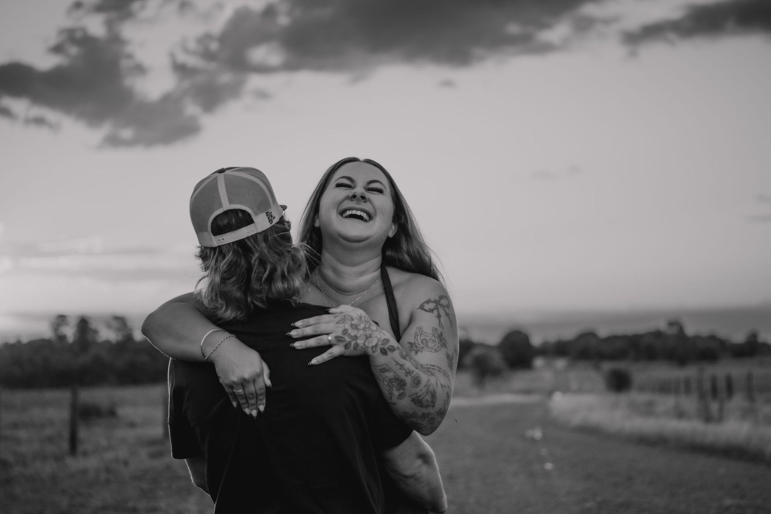 Black and white photo of a laughing tattooed couple embracing outdoors at sunset.