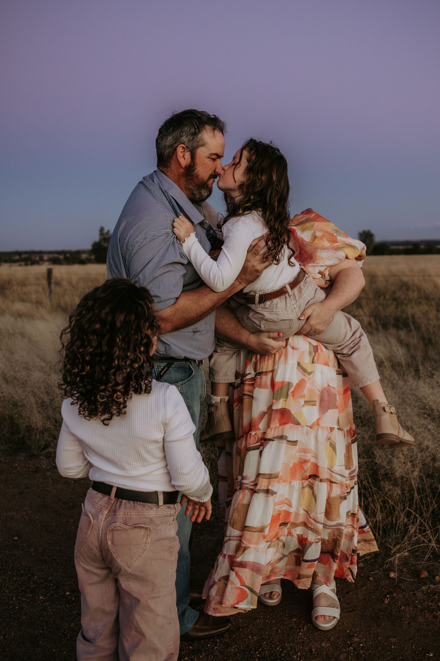A man kisses a woman he is holding while a curly-haired child watches, set against a golden field at dusk.