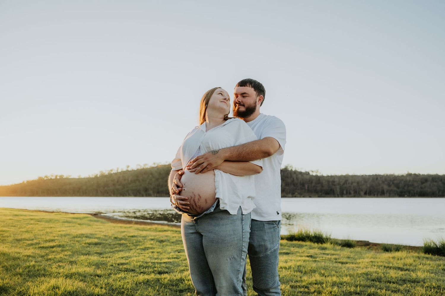 Couple embracing outdoors near a lake at sunset, pregnant woman smiling as man holds her baby bump.