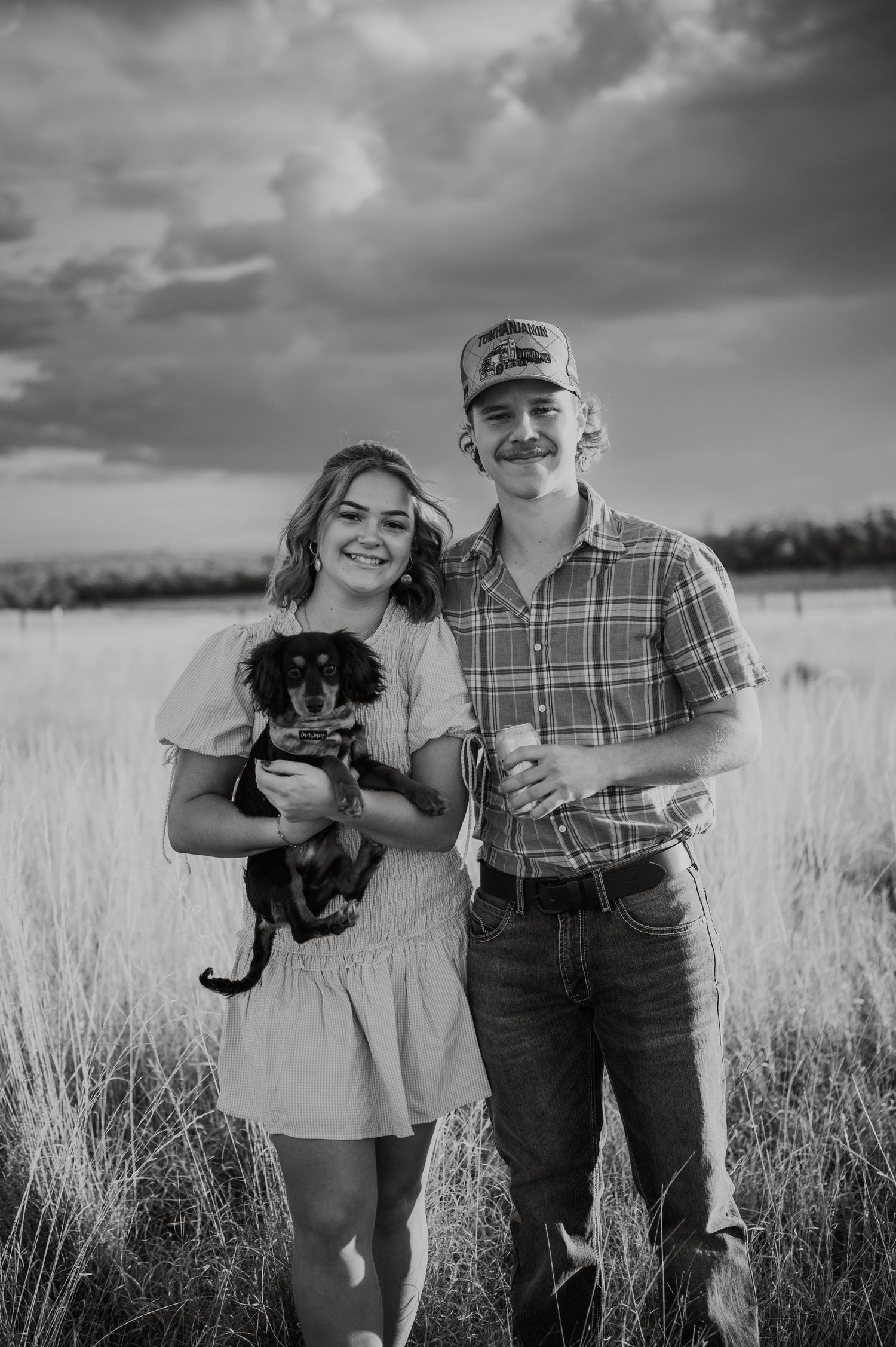 Black and white photo of a smiling couple standing in a field holding a dachshund dog under dramatic cloudy skies.
