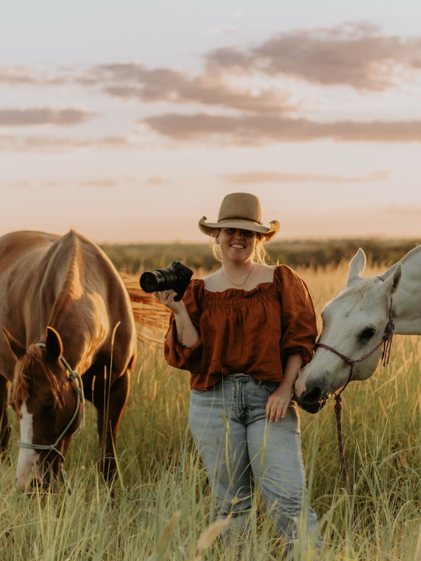 Person in cowboy hat with horses grazing in sunset-lit field.