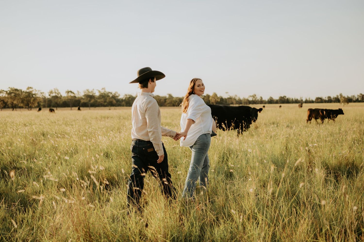 Couple walking through tall grass in a field with cattle grazing in the background.