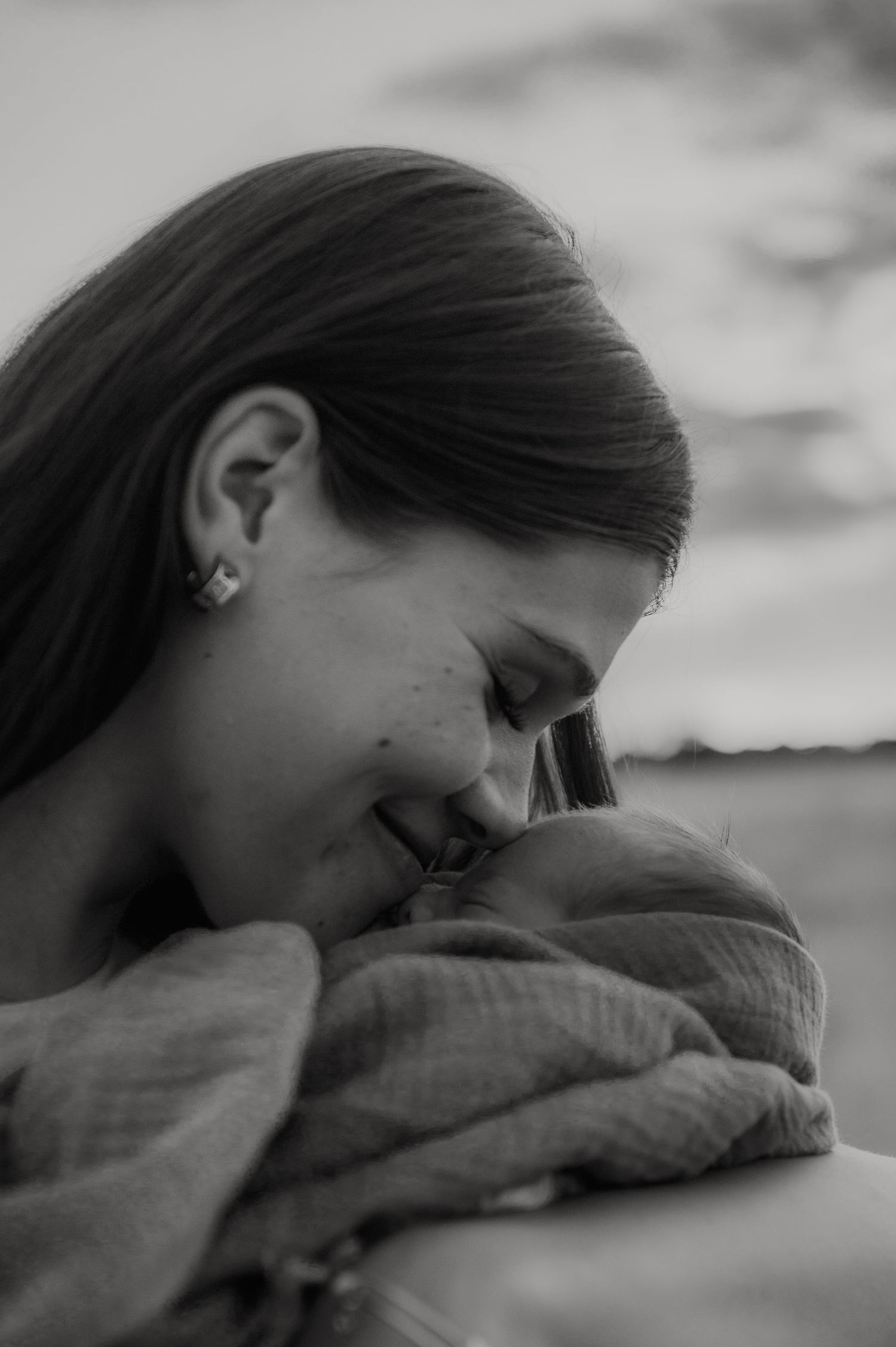 Black and white photo of a smiling mother tenderly nuzzling her newborn baby wrapped in a soft blanket.