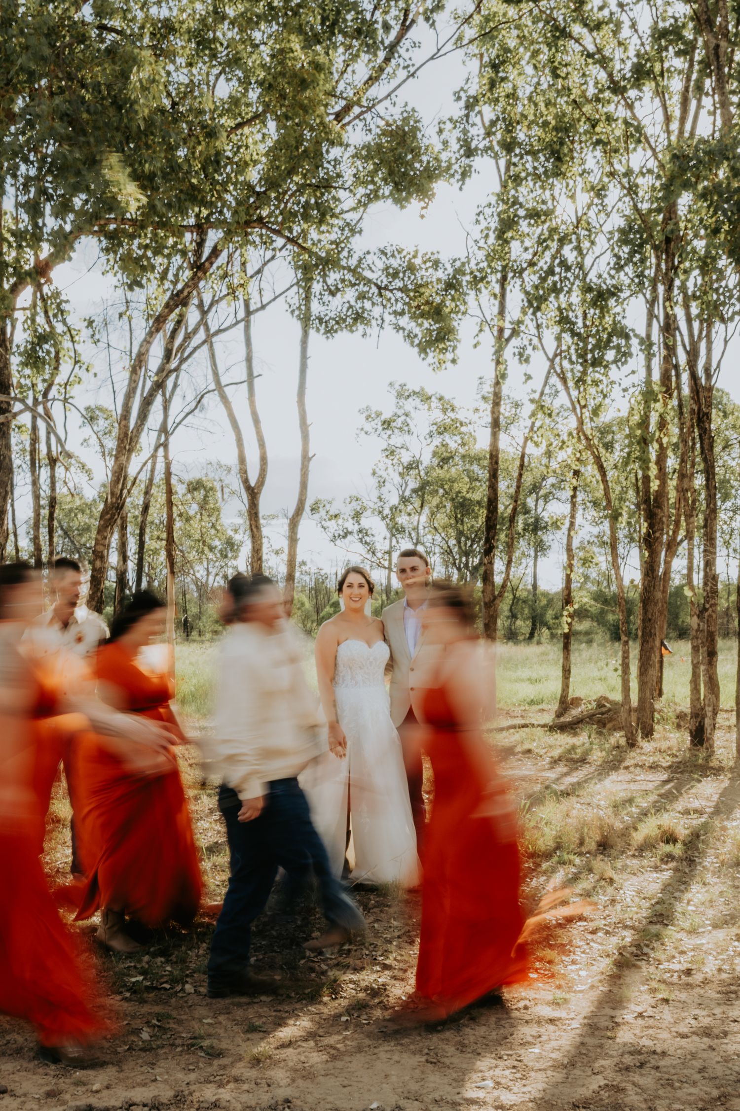 Bride and groom stand in sunlit forest with blurred bridesmaids in red dresses creating motion effect.