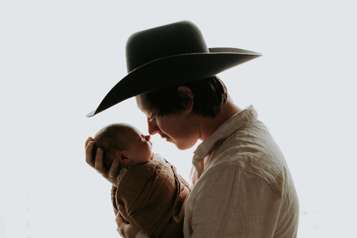 A parent wearing a cowboy hat tenderly holds and nuzzles a newborn baby in a soft, bright silhouette portrait.