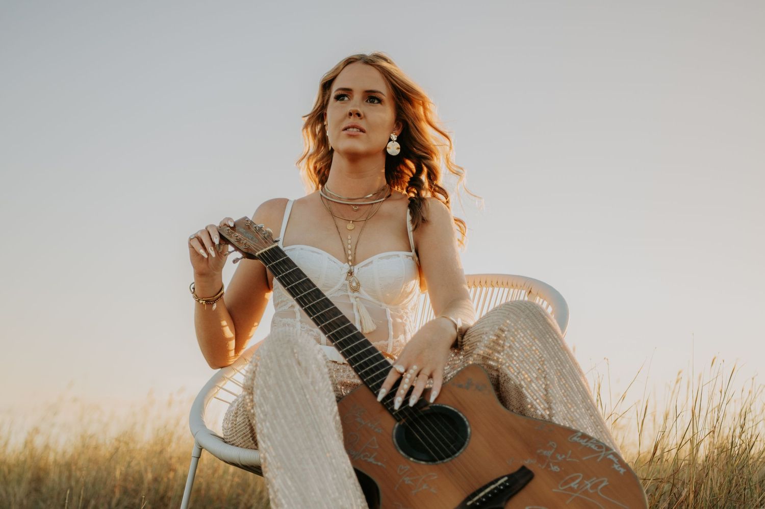 A woman in a white dress sits in a golden field with a guitar during sunset creating an ethereal outdoor portrait.