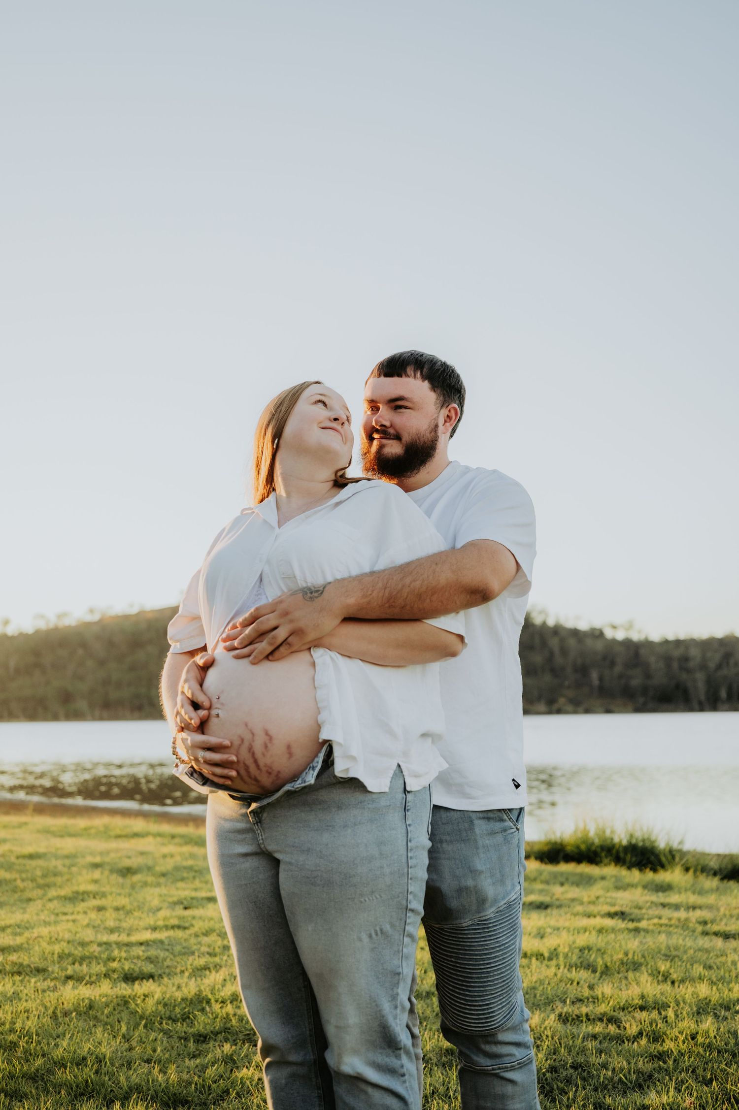 Couple sharing a tender moment outdoors by a lake, with the woman visibly pregnant, both smiling in golden hour light.
