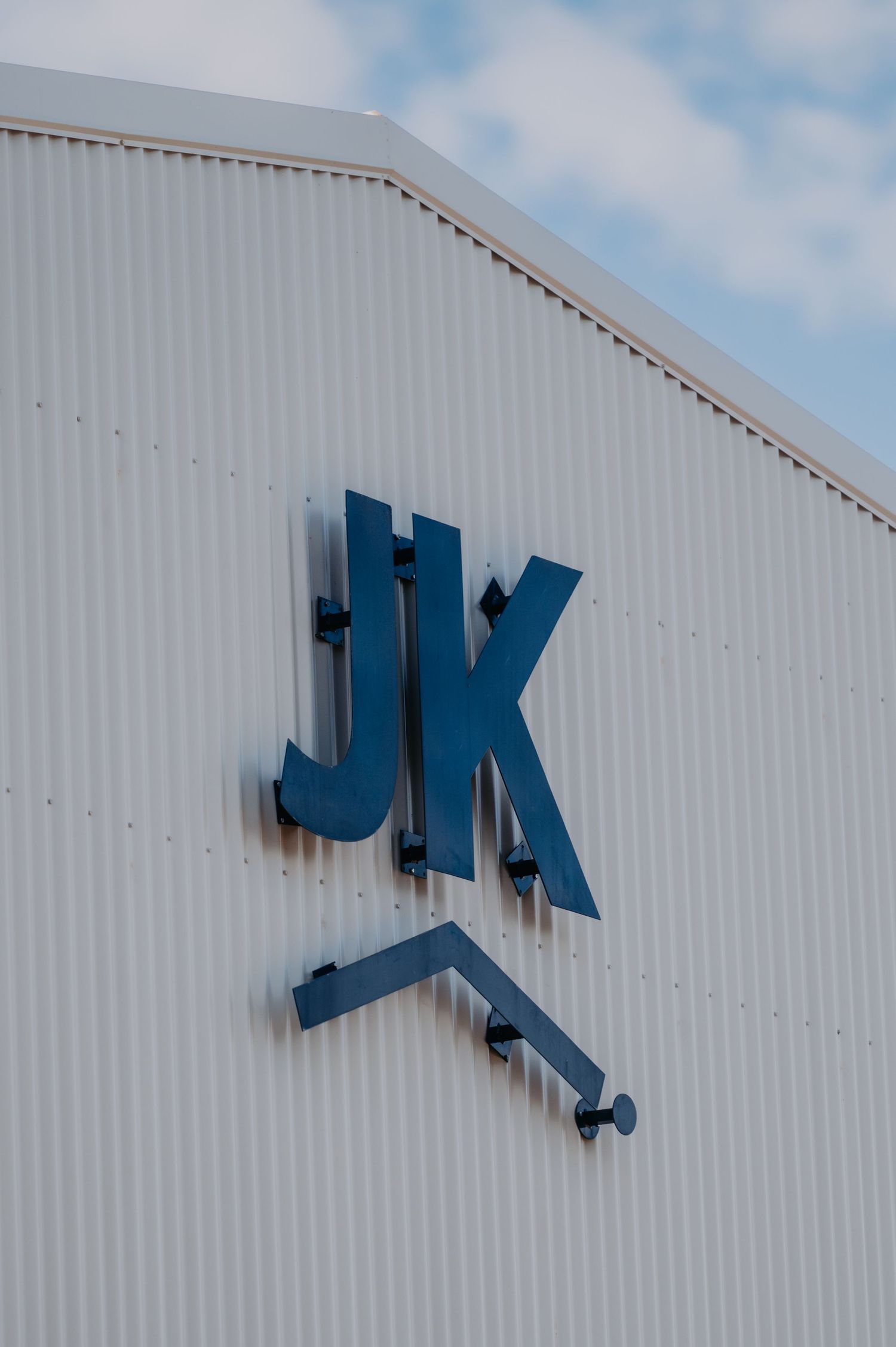 Blue JK logo sign mounted on a white corrugated metal building exterior against a cloudy sky.