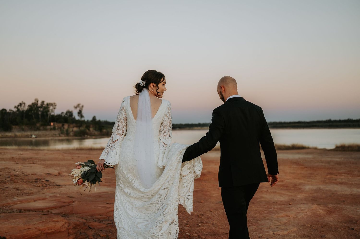 Bride and groom walk hand-in-hand at sunset near a lake, bride in lace gown holding bouquet.