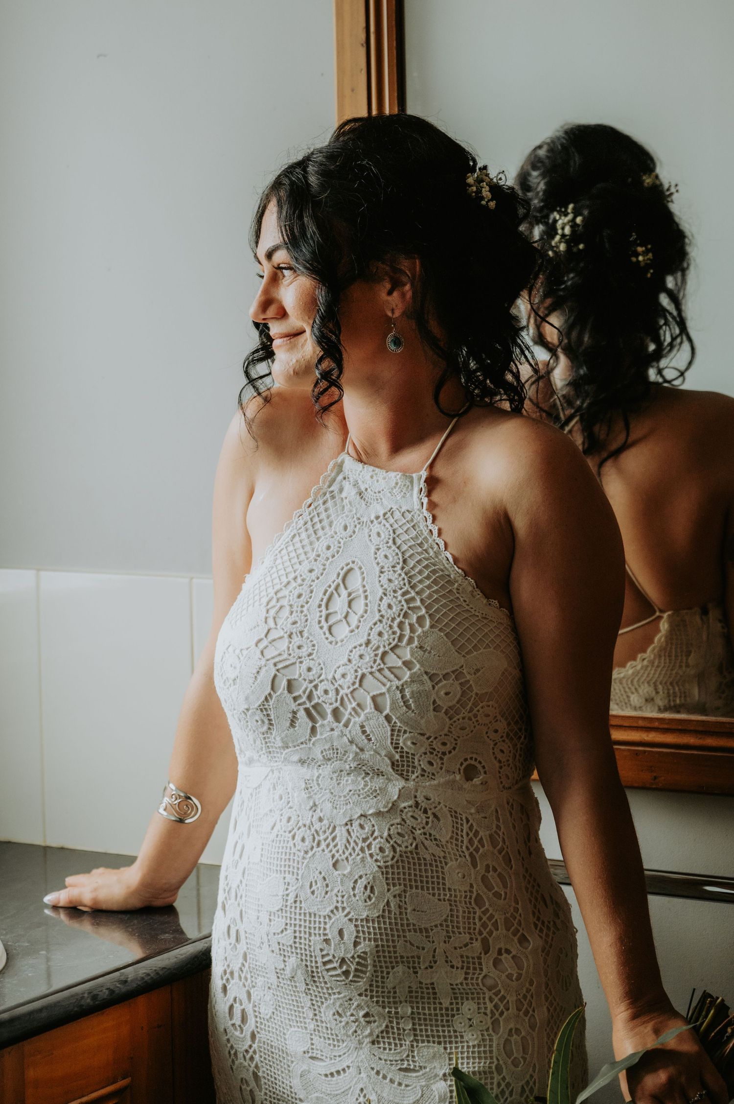 Bride in white lace halter wedding dress smiling near mirror, showing elegant back detail and floral hair accessories.