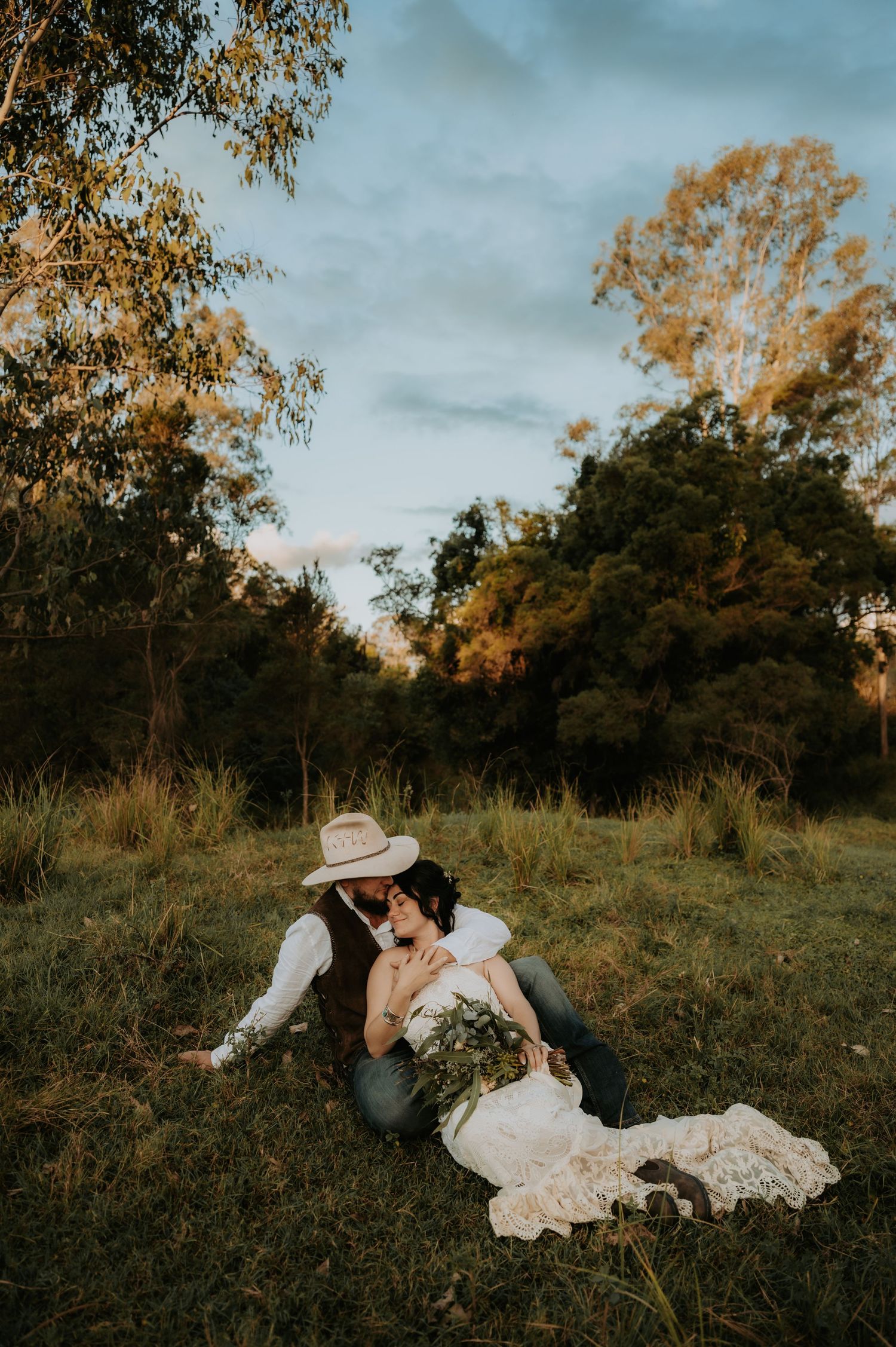 Couple sharing a romantic kiss in a rustic field at golden hour, bride in lace gown holding greenery bouquet.
