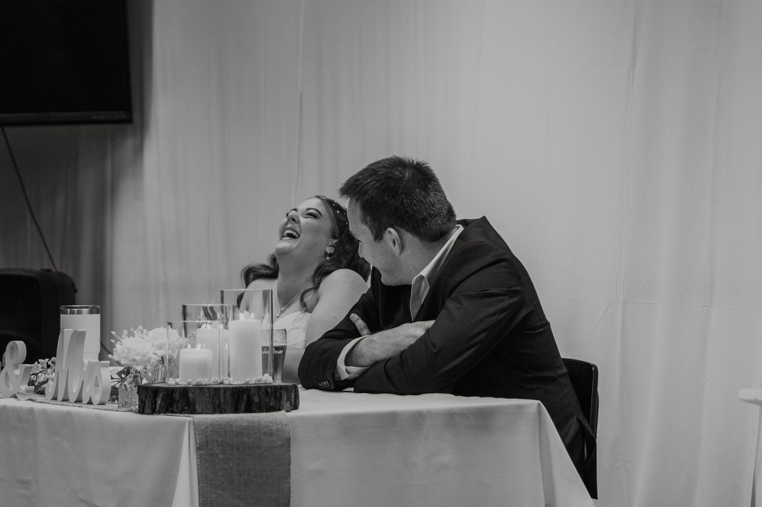 Black and white photo of bride and groom laughing joyfully at sweetheart table during wedding reception.