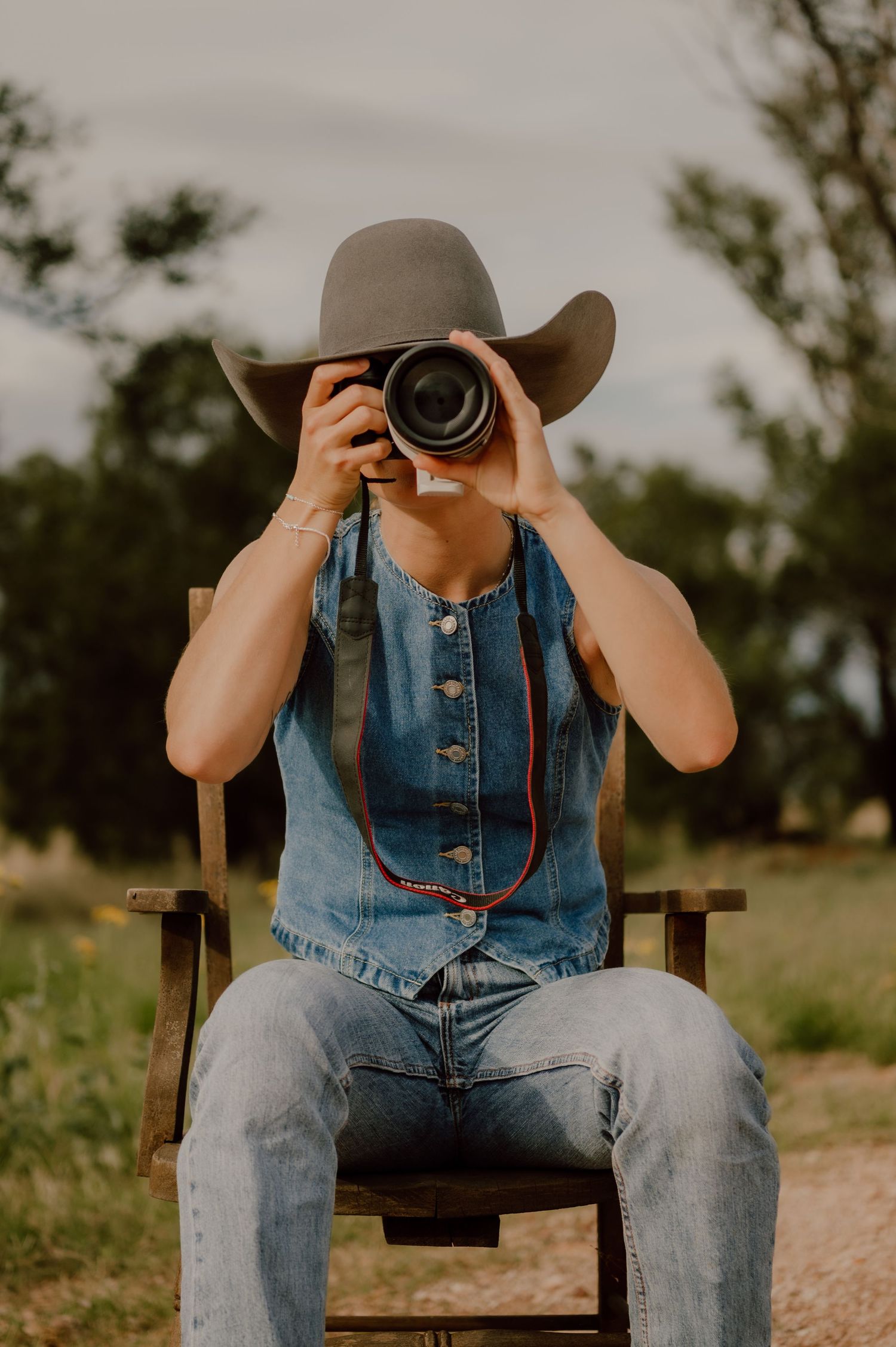 Photographer in denim overalls and cowboy hat sits on wooden chair taking photos in rustic outdoor setting.
