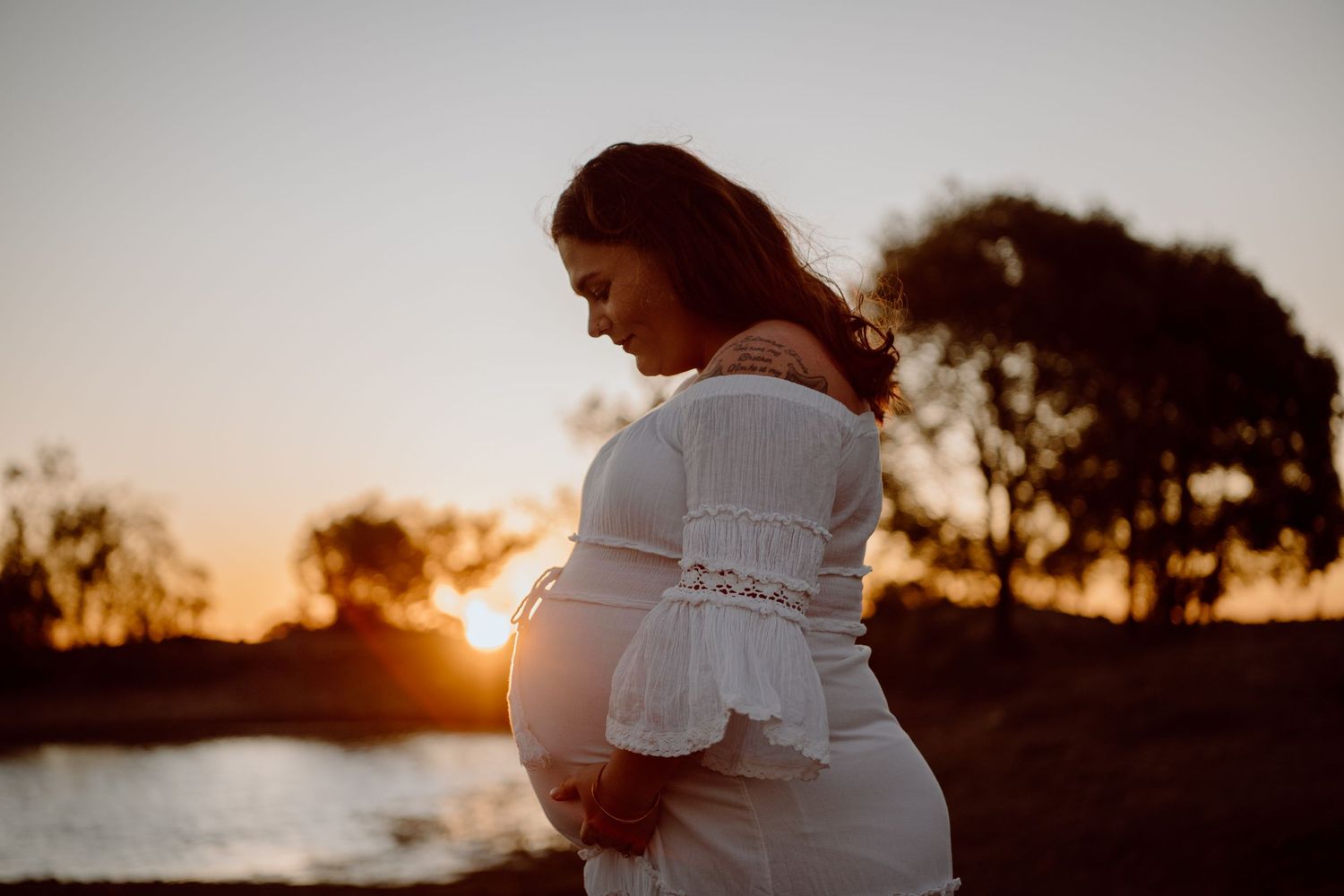 A side profile of a pregnant woman in white dress against a sunset waterfront.