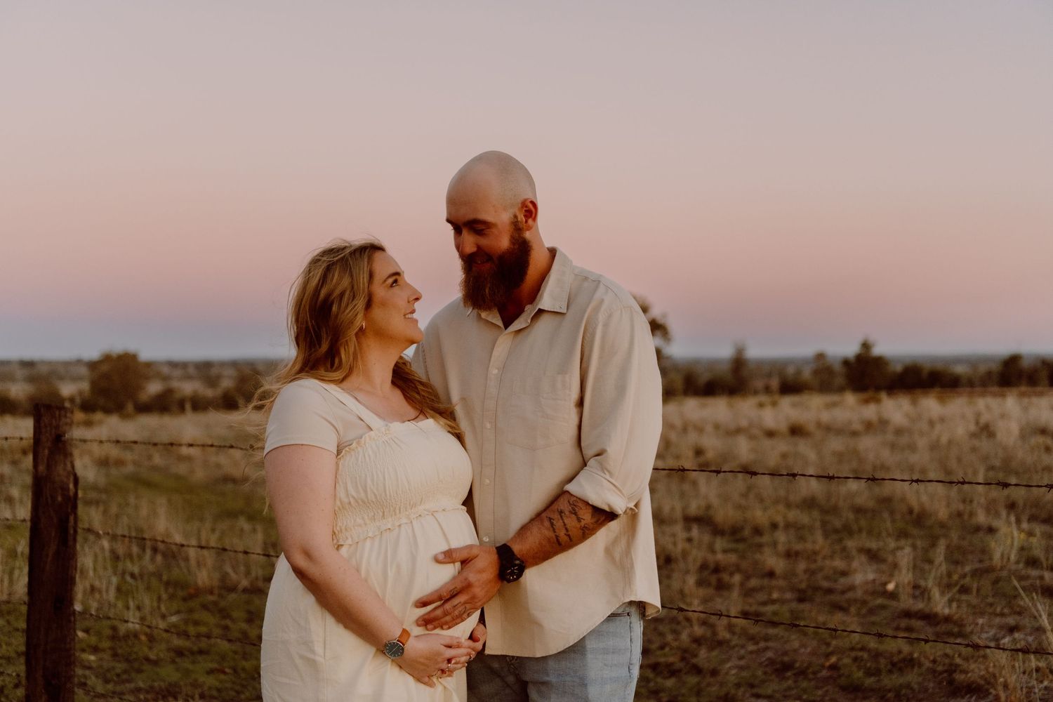 A couple shares an intimate moment at sunset in a rustic field setting during a maternity photoshoot.