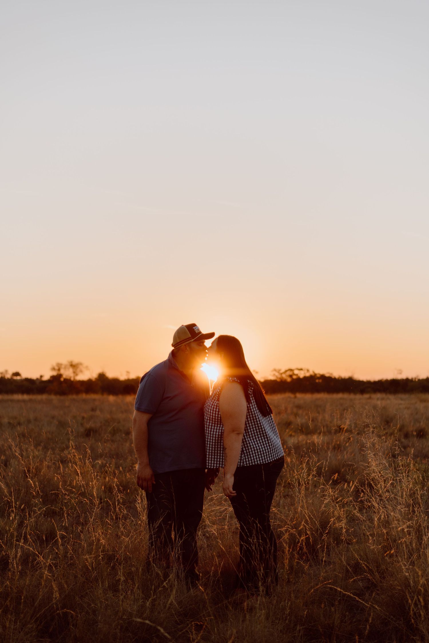 Couple sharing a romantic kiss silhouetted against a golden sunset in an open field.