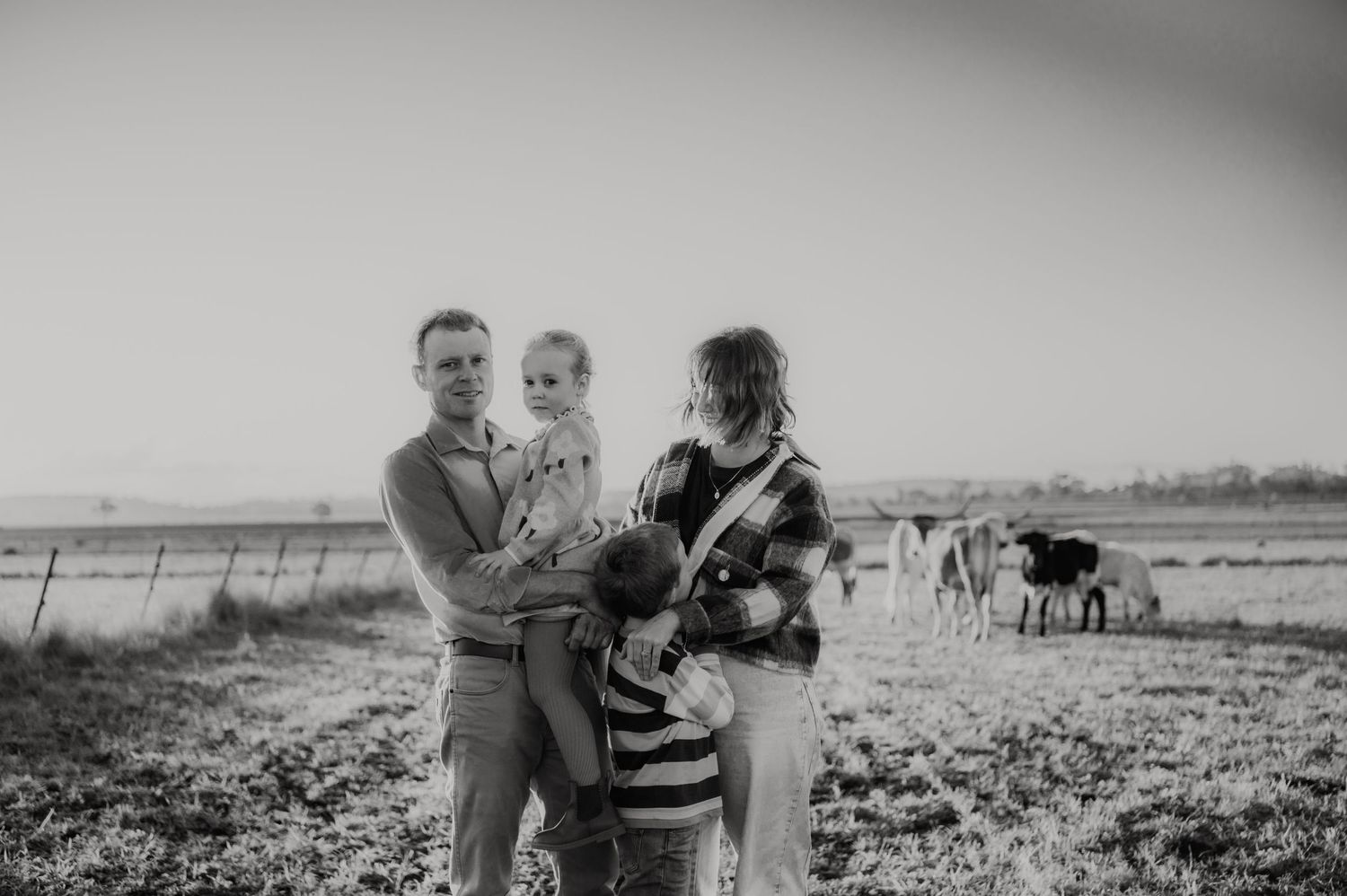 Black and white family photo with parents holding children in a farm field with cattle grazing in the background.