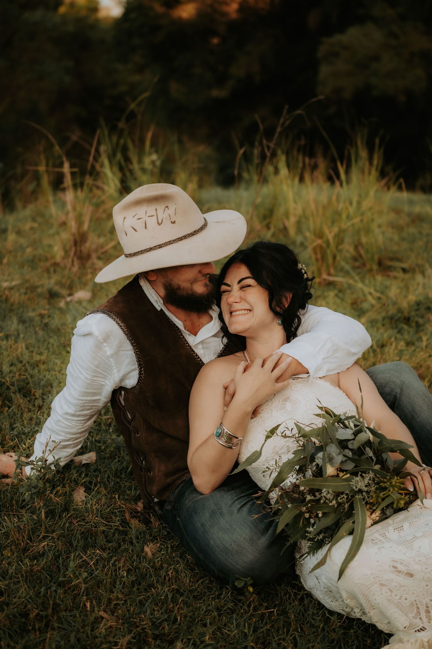 A couple in rustic wedding attire sits on grass, the groom in a cowboy hat nuzzling the smiling bride holding a greenery bouquet.