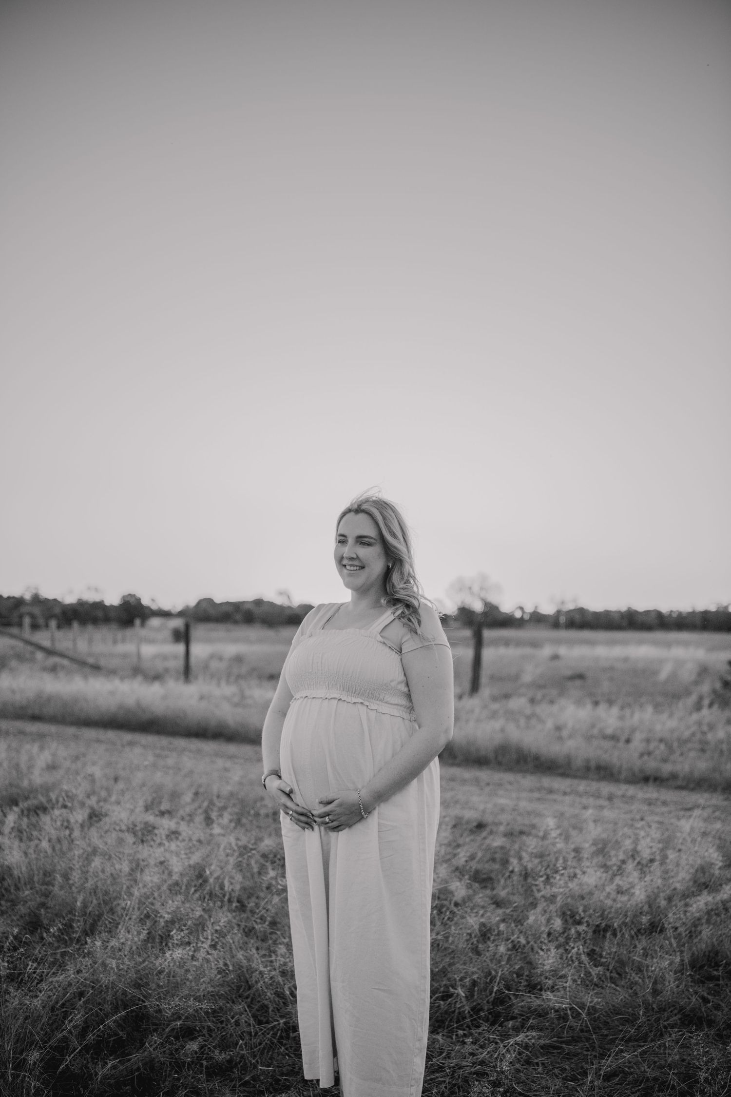 A pregnant woman in a light maxi dress stands in a field during a black and white maternity photoshoot.