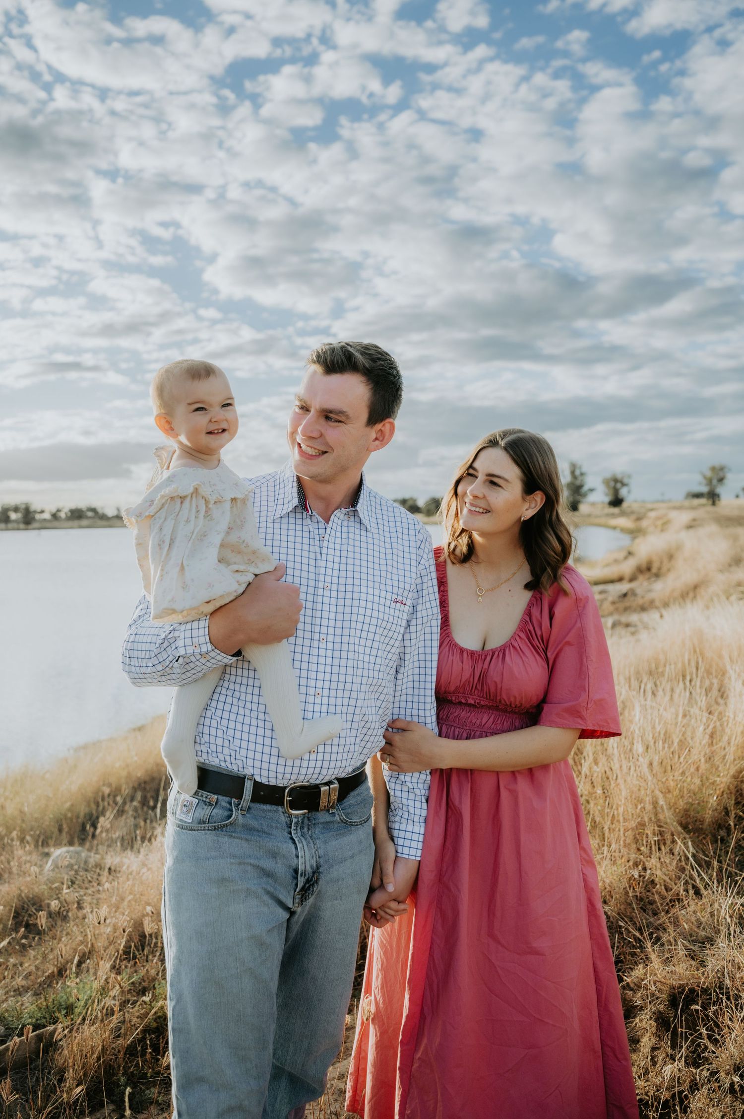 A happy young family with a baby poses outdoors near a lake in a golden field under a cloudy sky.