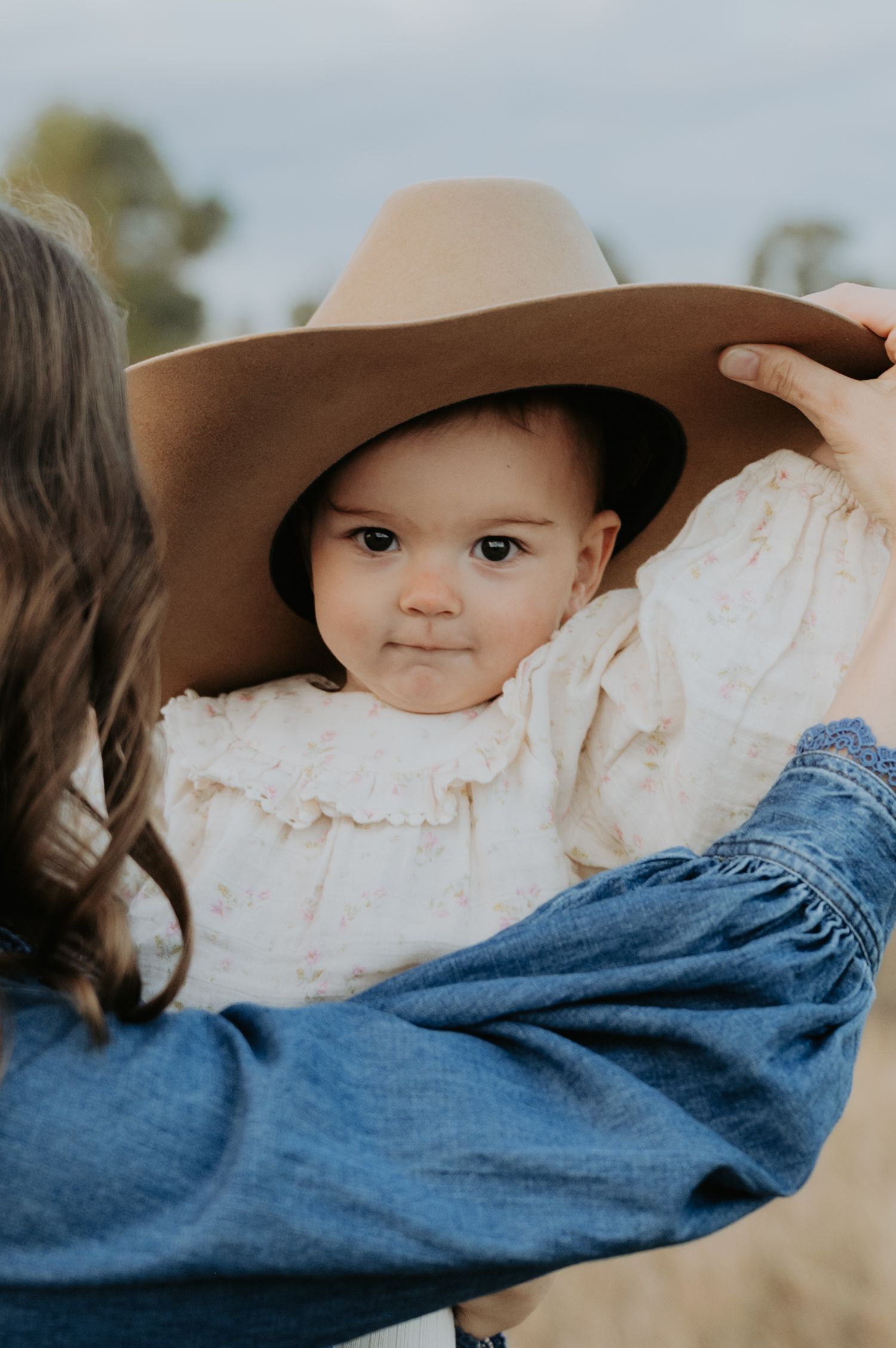 A woman in a denim shirt places a tan cowboy hat on an adorable baby wearing a white floral dress outdoors.