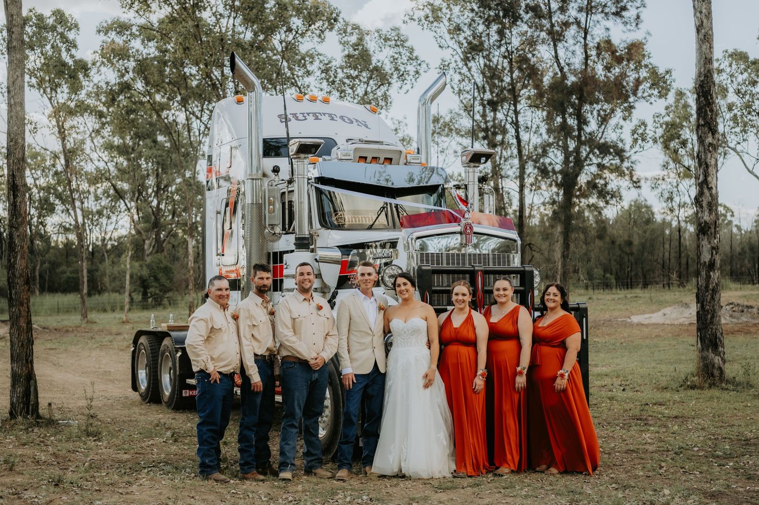 Wedding party poses in front of a large semi-truck outdoors, with bridesmaids in red dresses and groomsmen in tan suits.