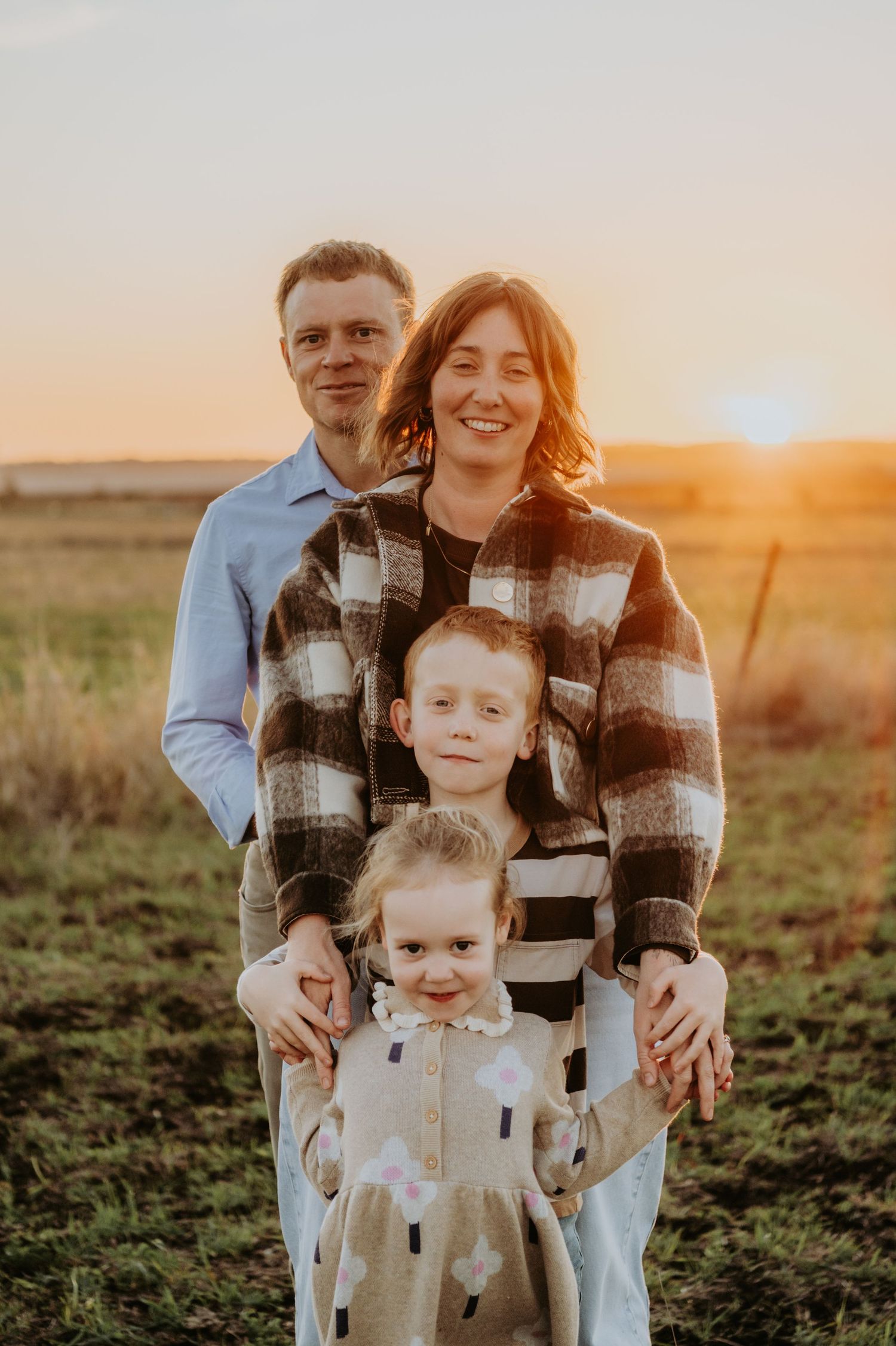 Family of four posing at golden hour in a field, parents standing behind two young children smiling at camera.