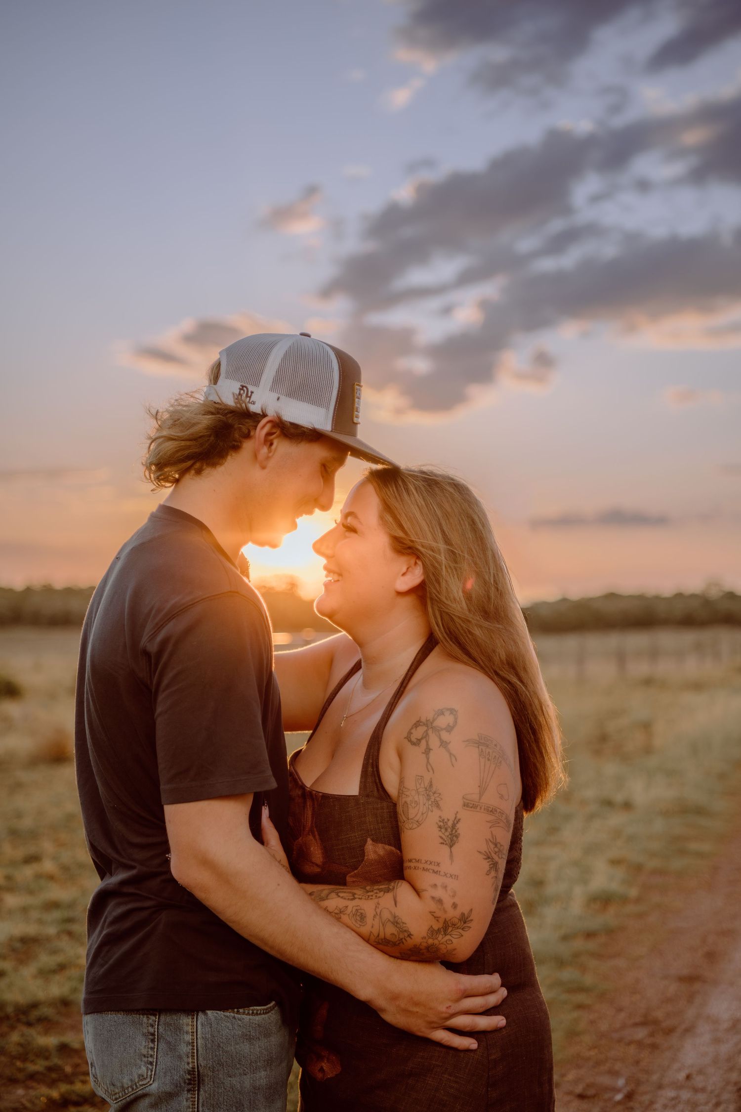 Couple embracing and almost kissing at golden sunset in open field, woman with tattoos wearing overalls, man in trucker hat.