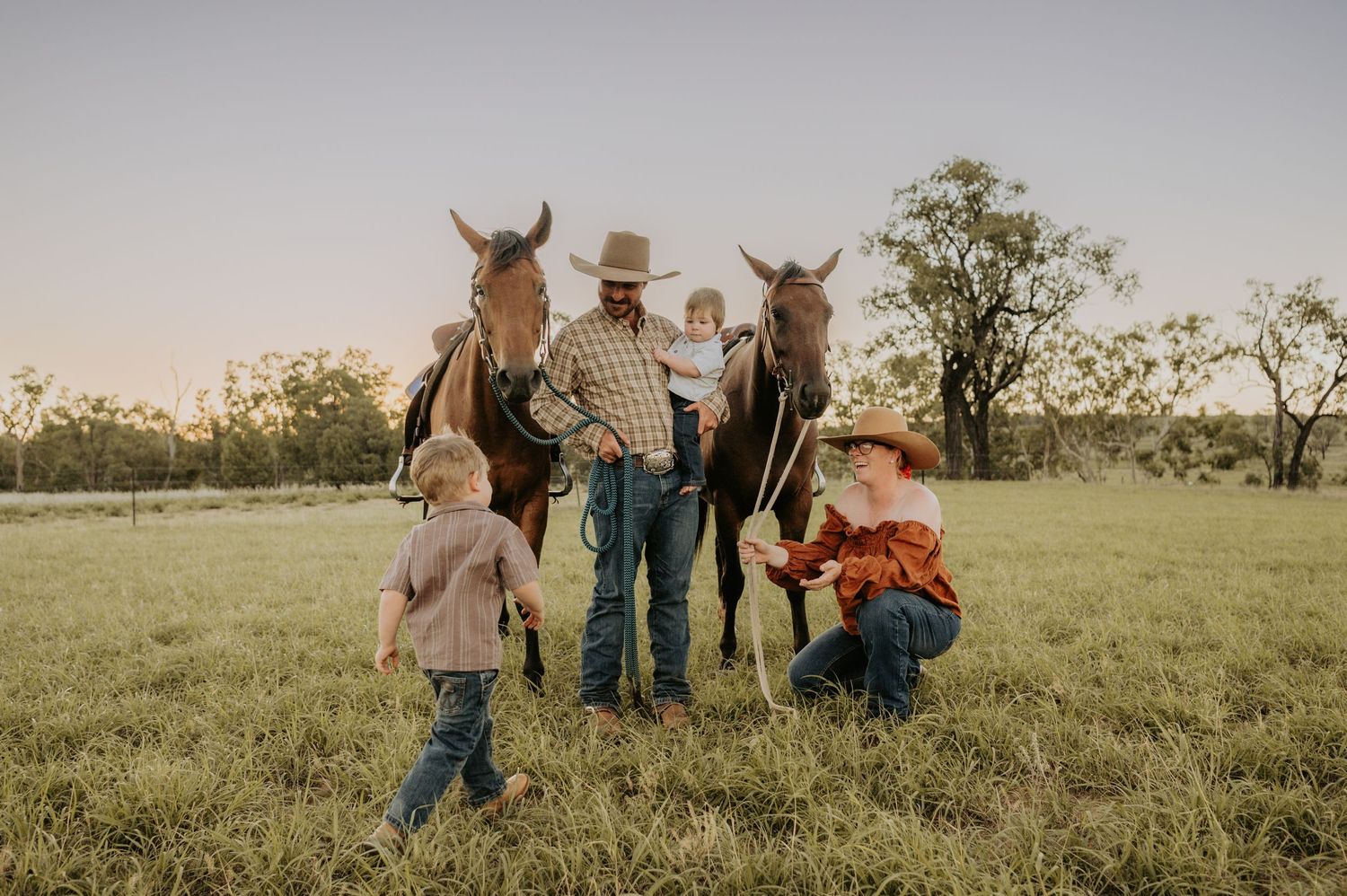 Family with two children and two horses in a green pasture at sunset, wearing cowboy hats and western attire.