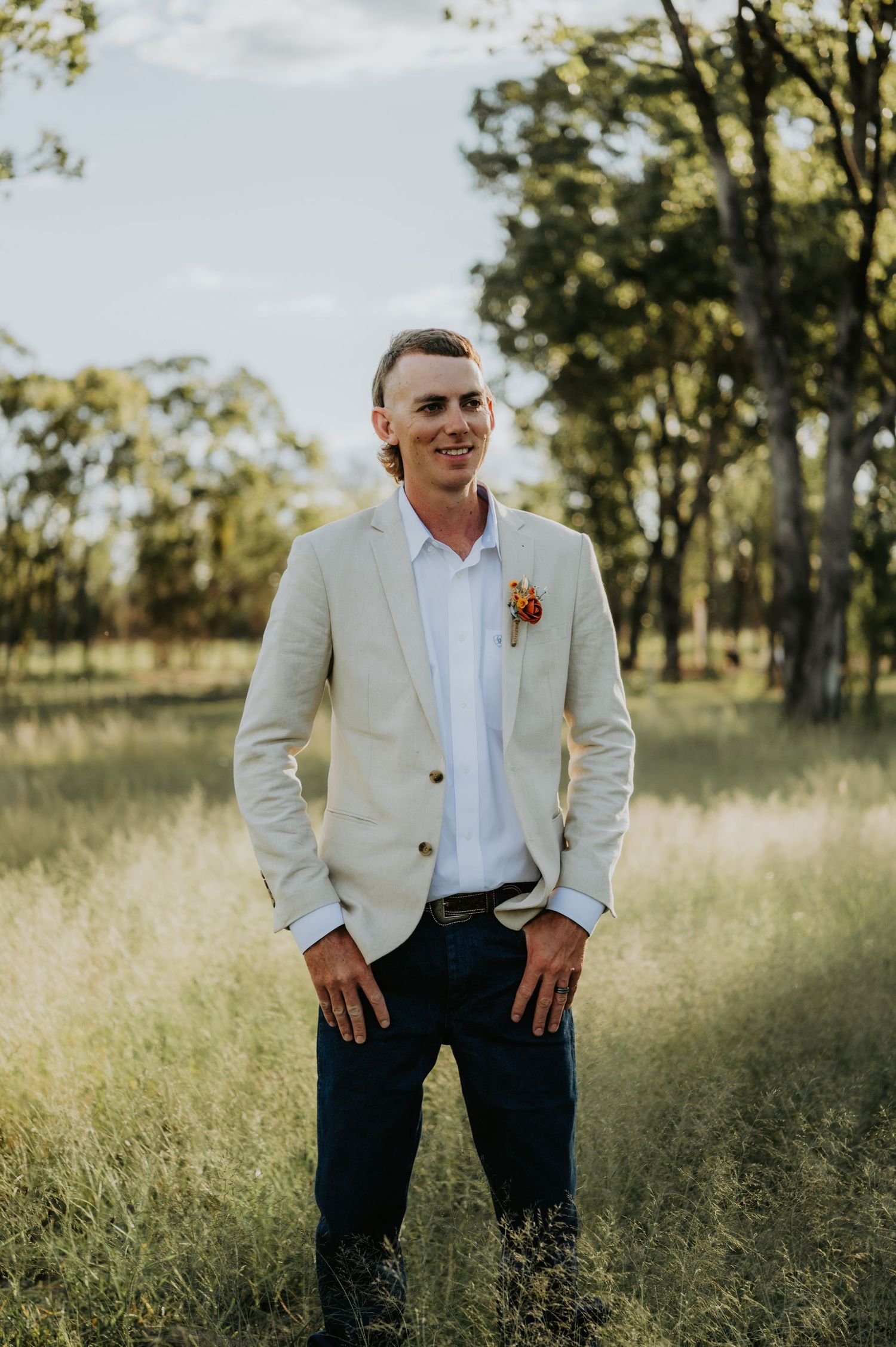Smiling groom in cream blazer with boutonniere stands in lush green field with trees in background.