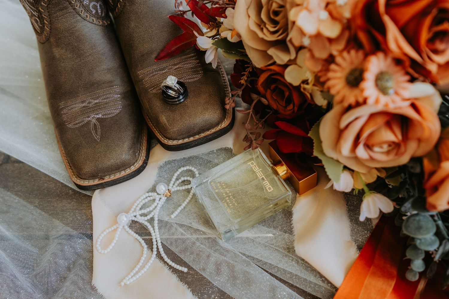 Rustic wedding flat lay with cowboy boots, rings, perfume, garter, and a bouquet of peach and orange roses.