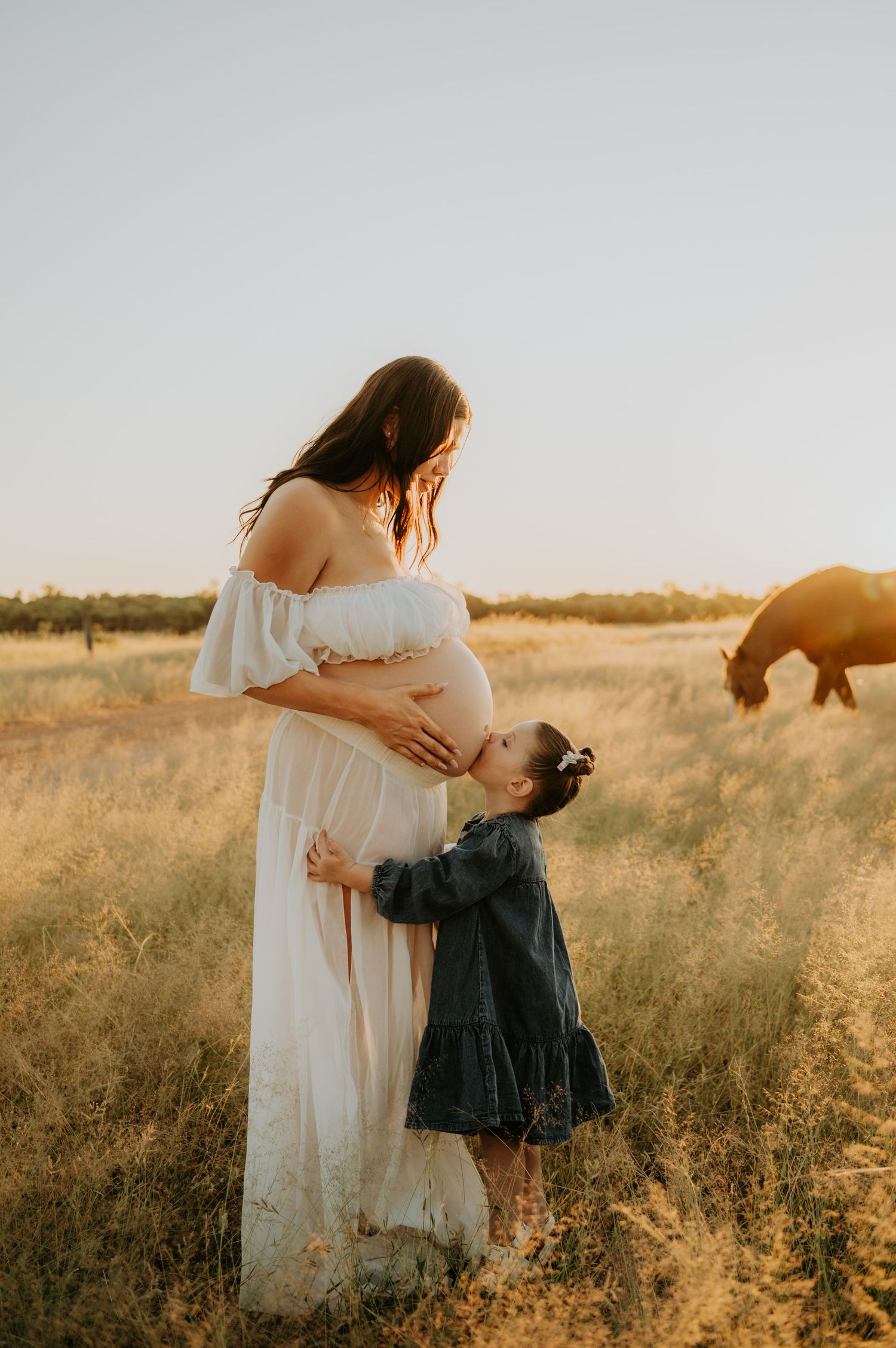 A young girl kisses a pregnant woman's belly in a golden field at sunset, with a horse grazing nearby.