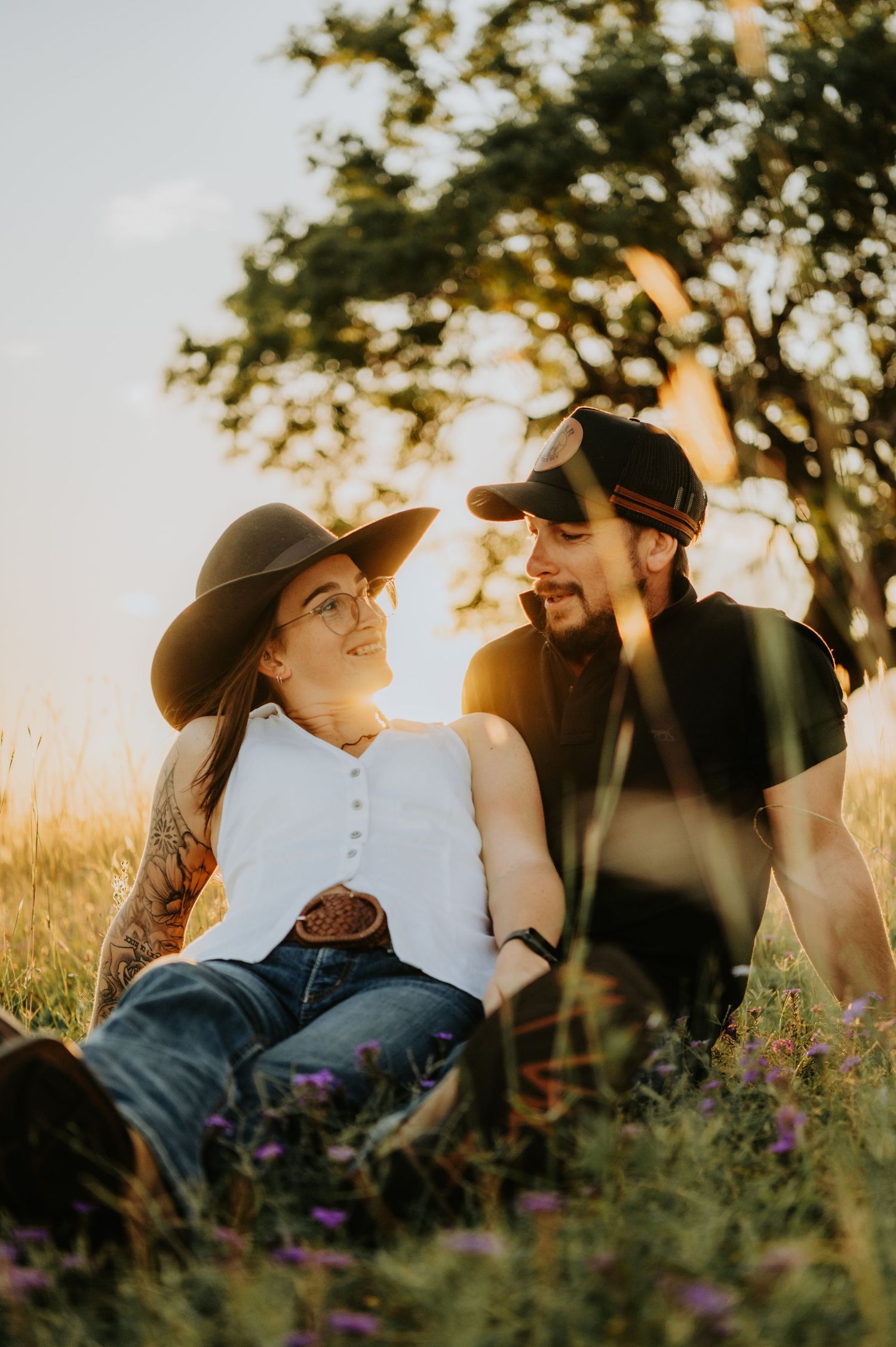 Couple sitting in golden hour sunlight in a grassy field, woman in cowboy hat and jeans smiling at tattooed man in black cap.