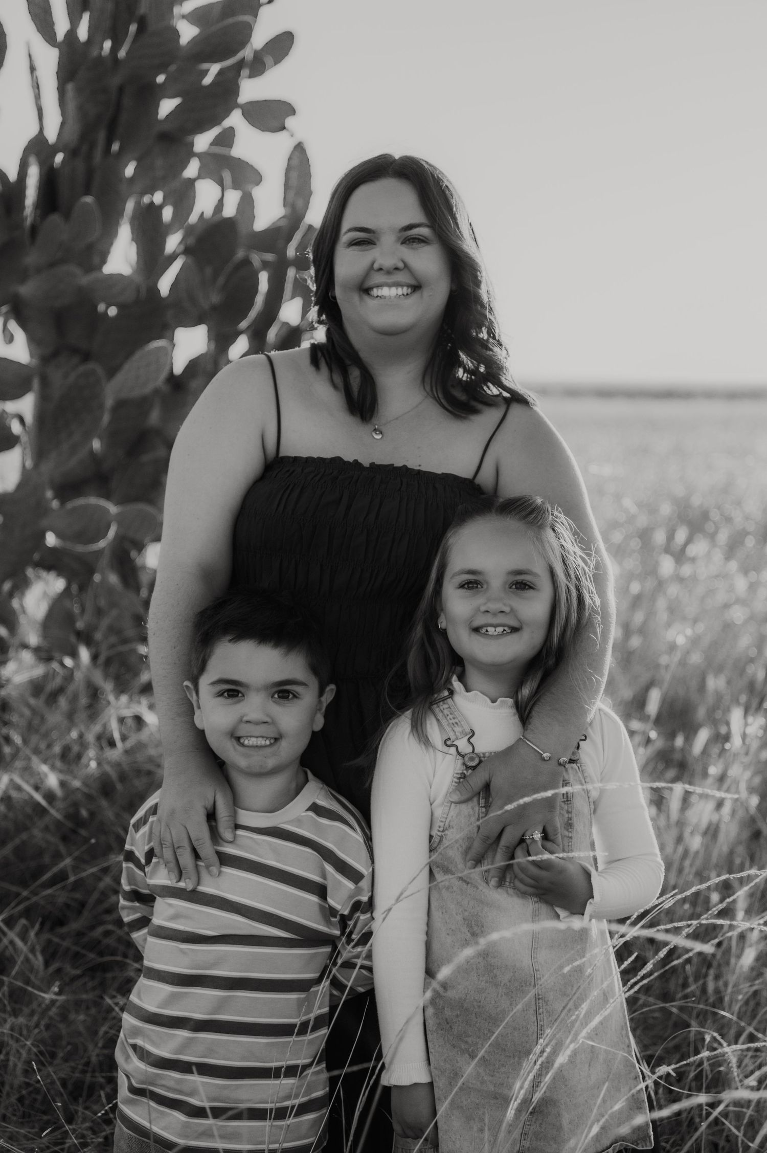 Black and white photo of a smiling woman with two young children outdoors near a cactus in a field.