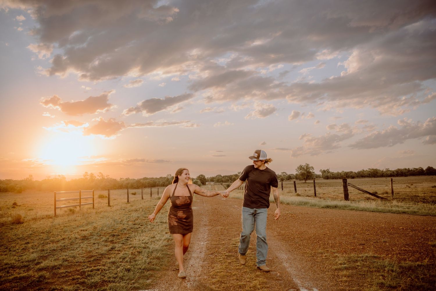Panoramic sunset view of couple walking through golden field with fence in background.