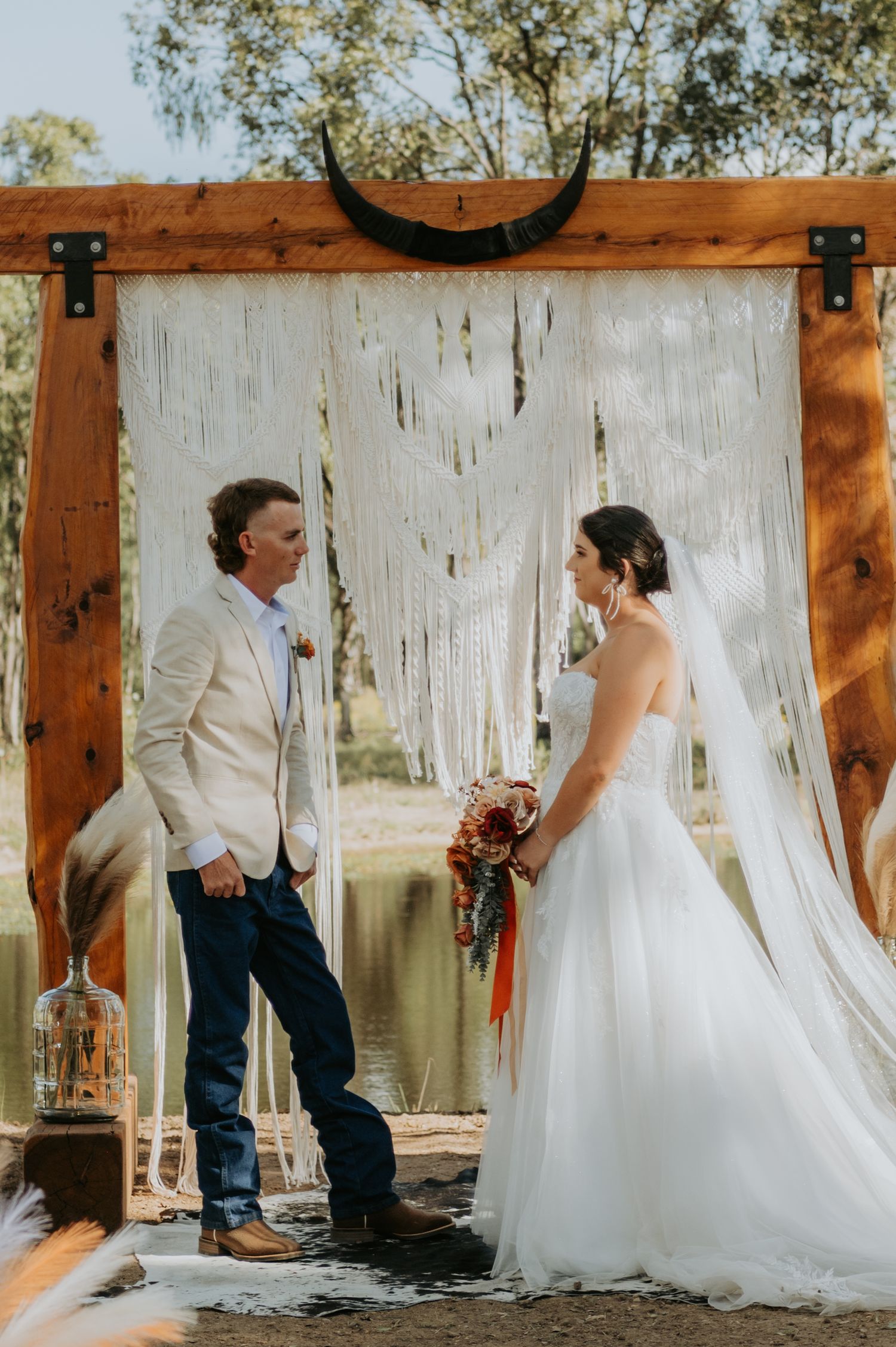 Bride and groom exchange vows under rustic wooden arch with macrame backdrop near serene pond at boho outdoor wedding.