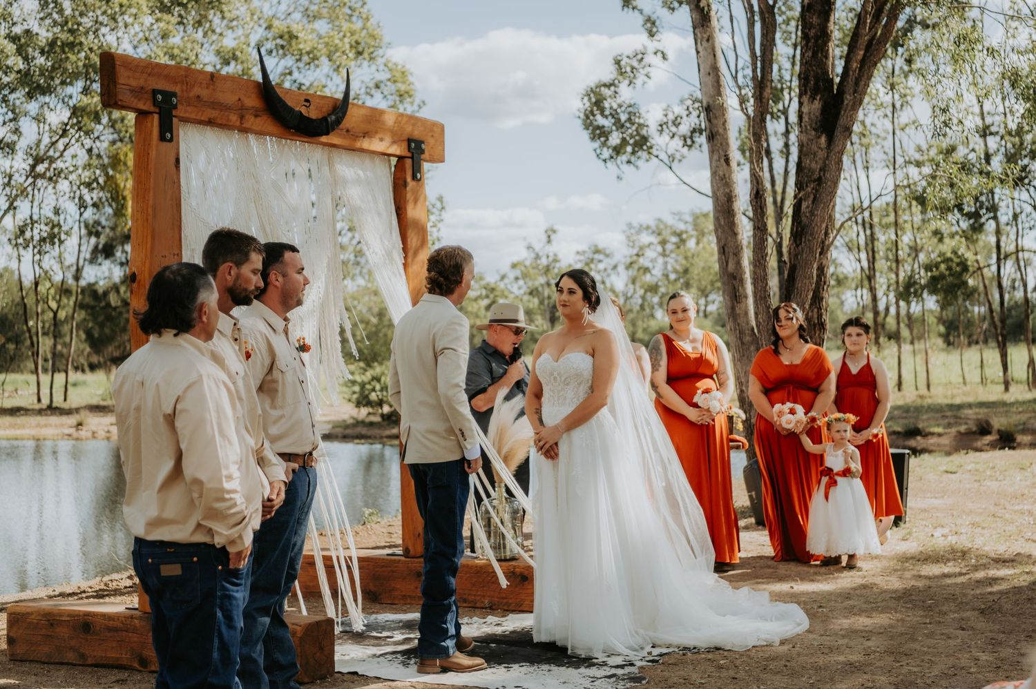 Outdoor rustic wedding ceremony by a pond with bride, groom, officiant, groomsmen, and bridesmaids in red dresses.