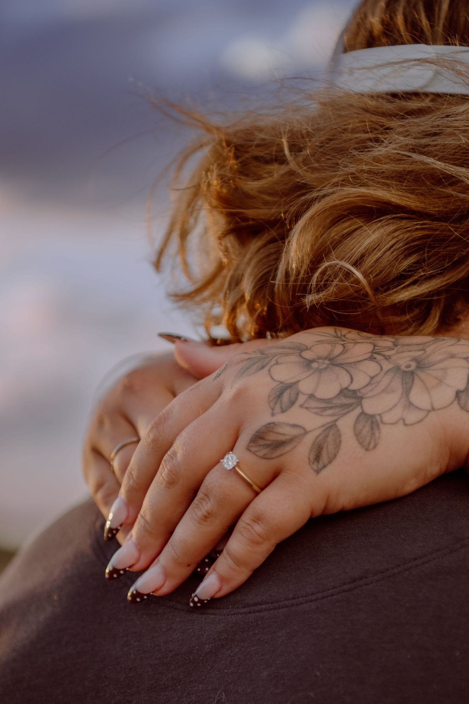 Close-up of a woman's hand with an engagement ring and floral tattoo resting on a man's shoulder.