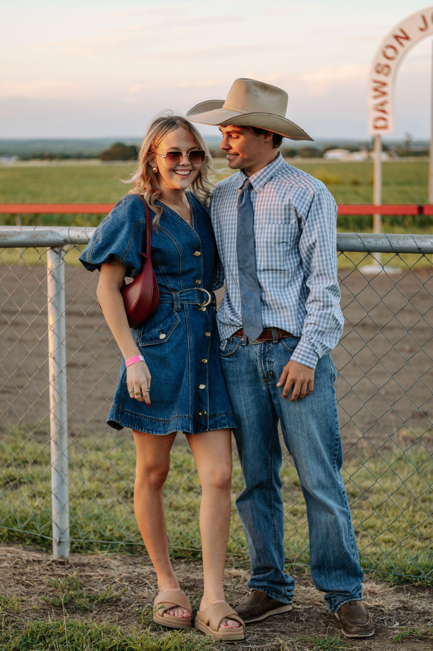 Couple in western attire posing at a rodeo, woman in denim dress, man in plaid shirt and cowboy hat.