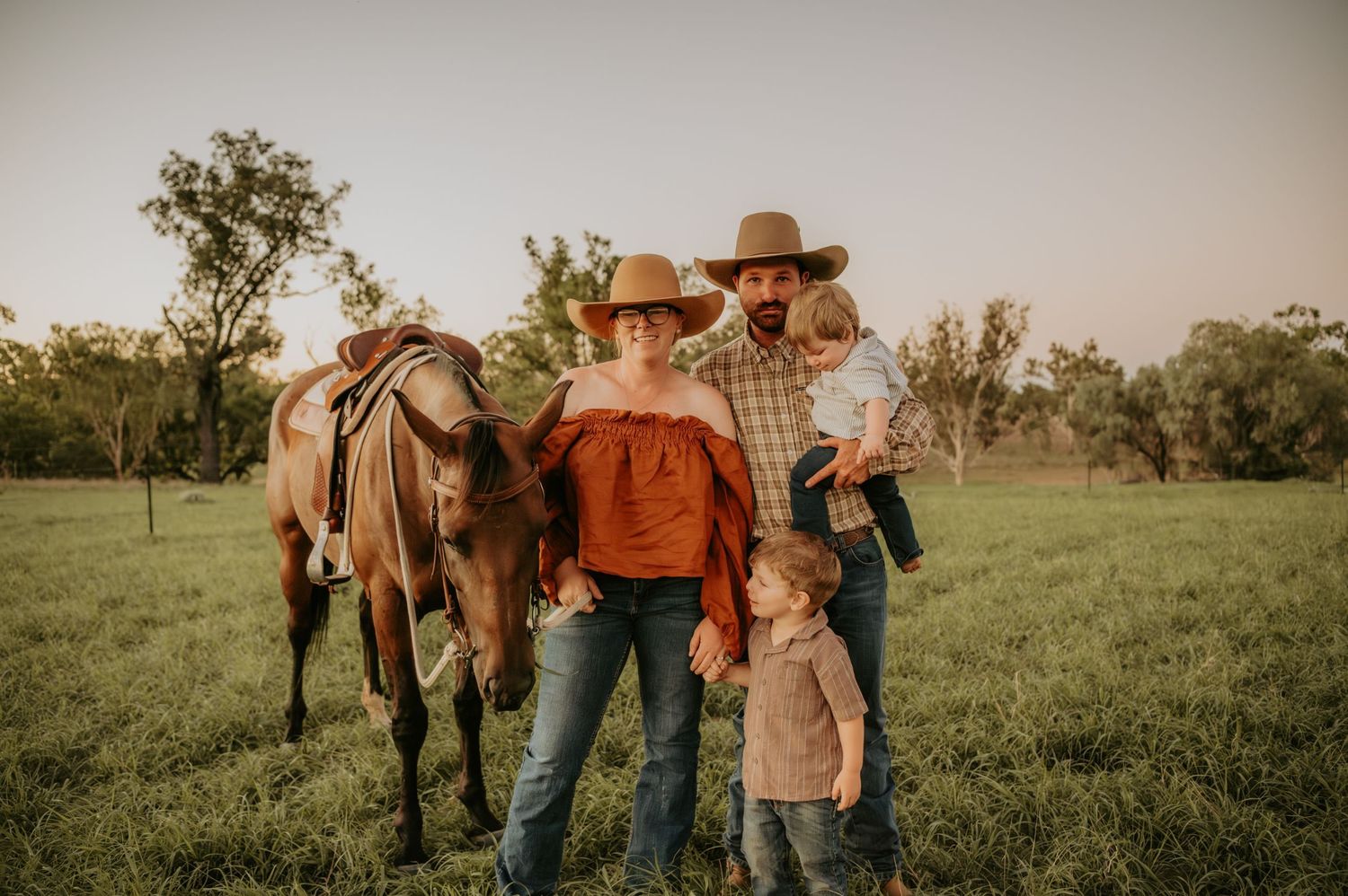 Family in cowboy hats posing with a brown horse in a green field at sunset.