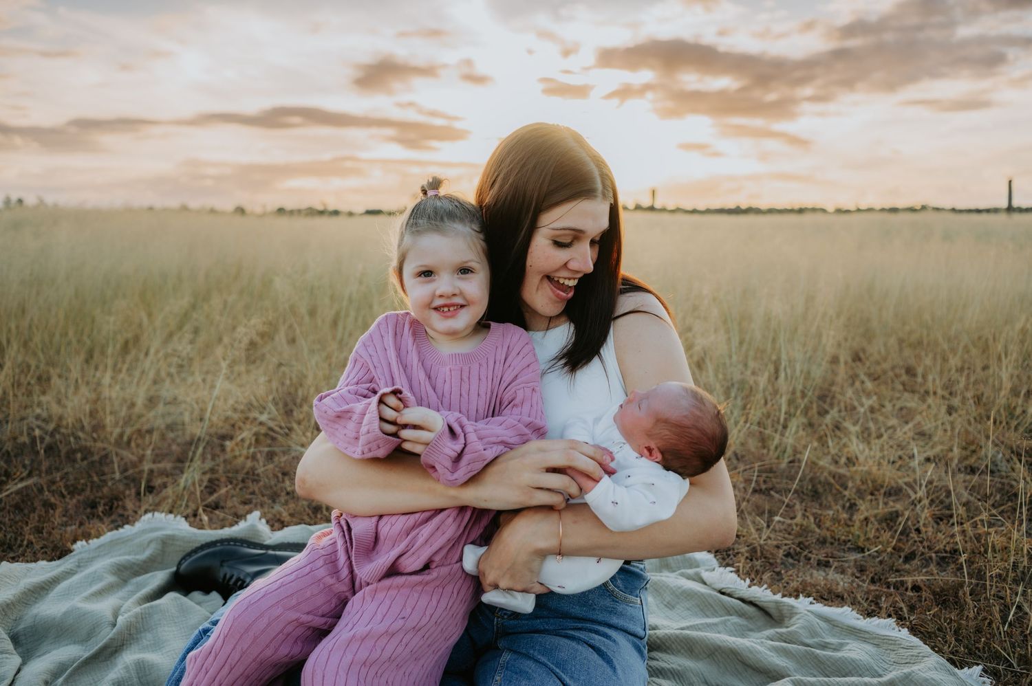 Mother sitting in a golden field at sunset, smiling while holding a newborn and a young girl in pink.