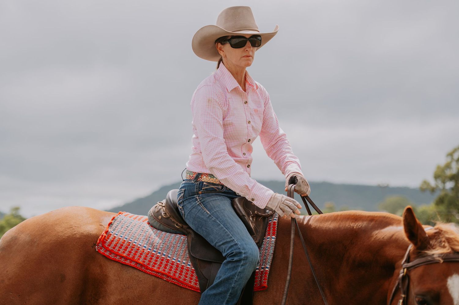 Woman in pink shirt and cowboy hat riding a chestnut horse in an outdoor western setting.