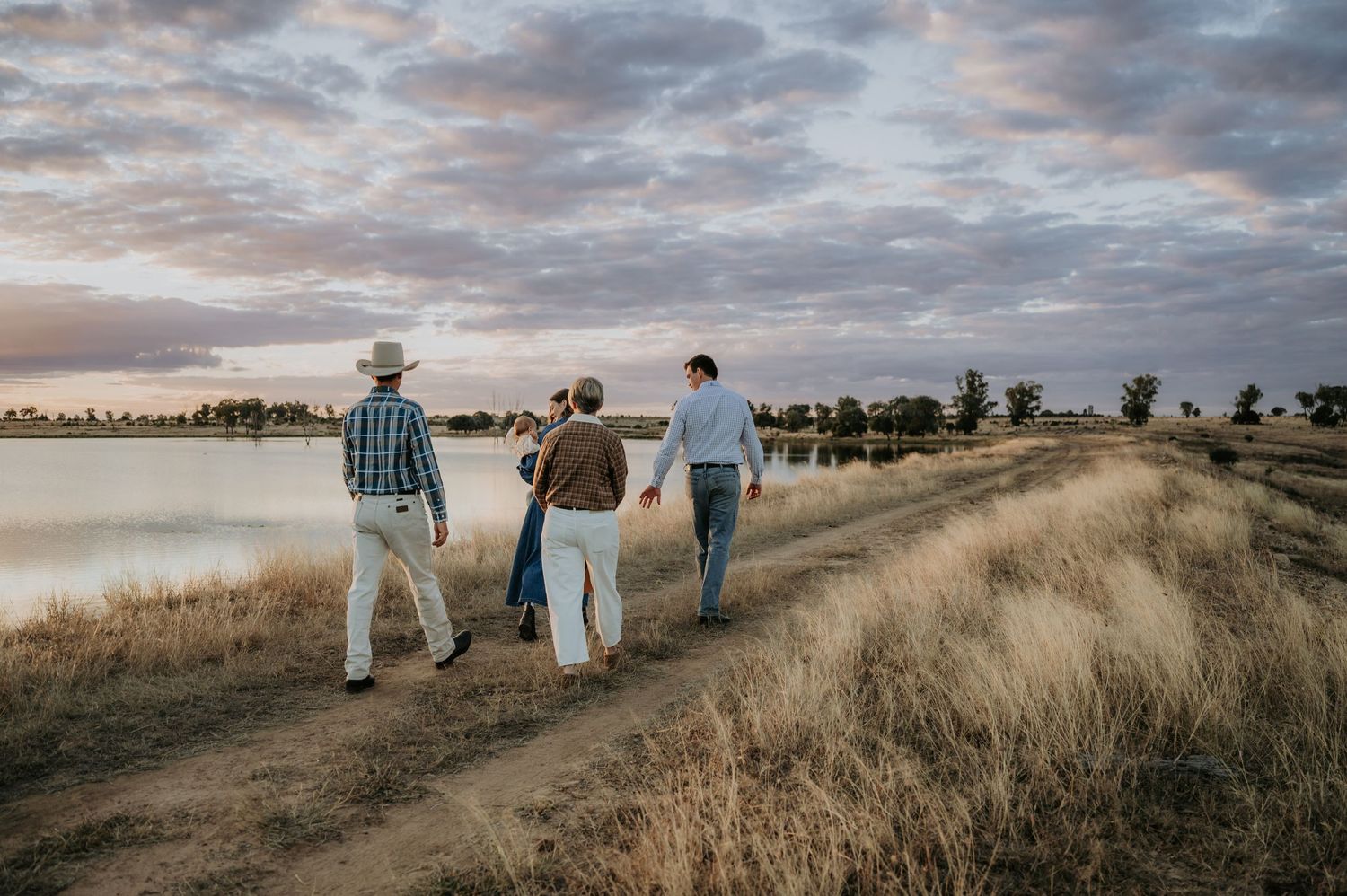Three people walking along a dirt path beside a calm lake at sunset in a rural countryside landscape.