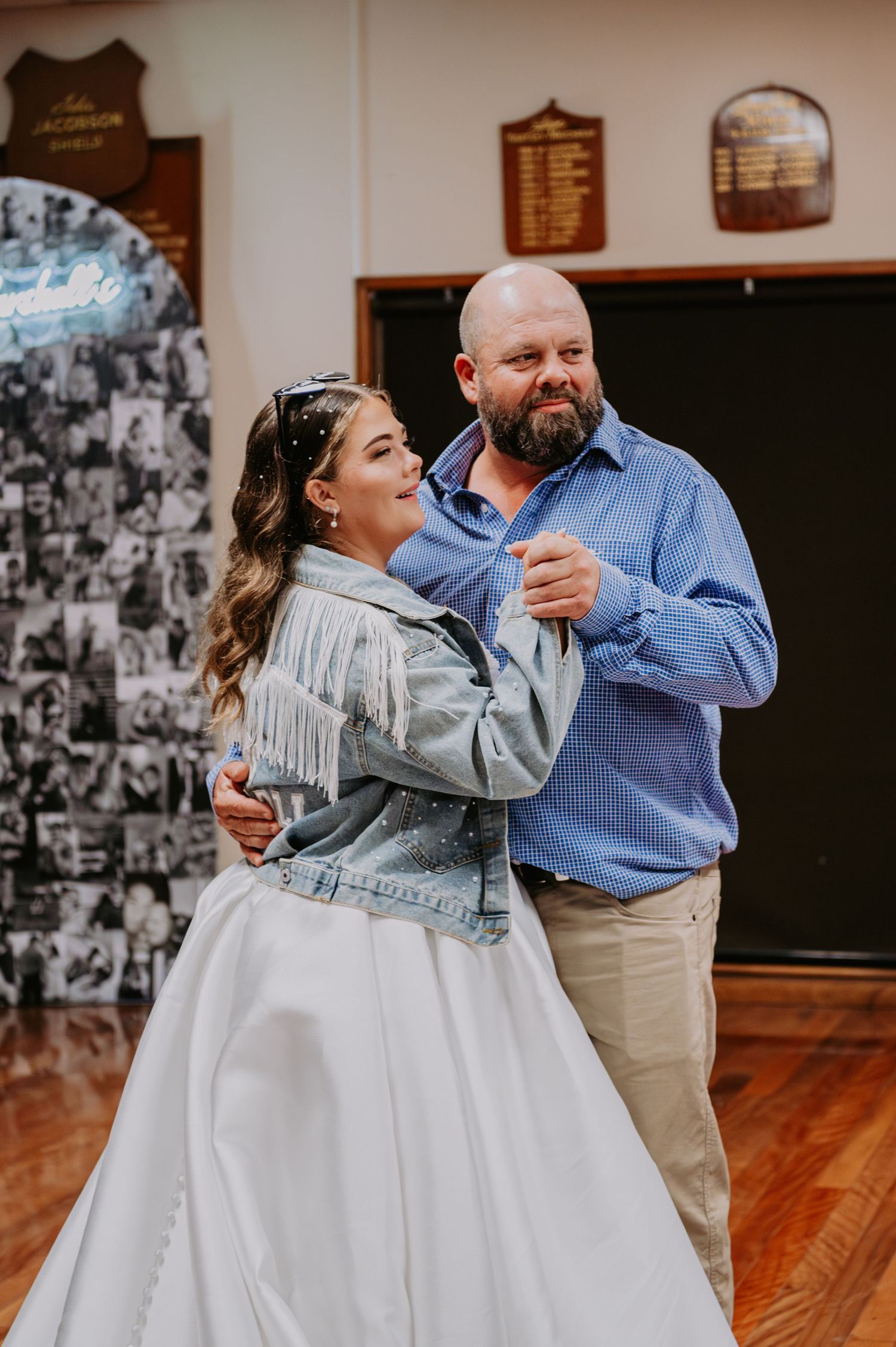 Bride in white gown and denim fringe jacket shares first dance with bald bearded man in blue shirt at wedding reception.