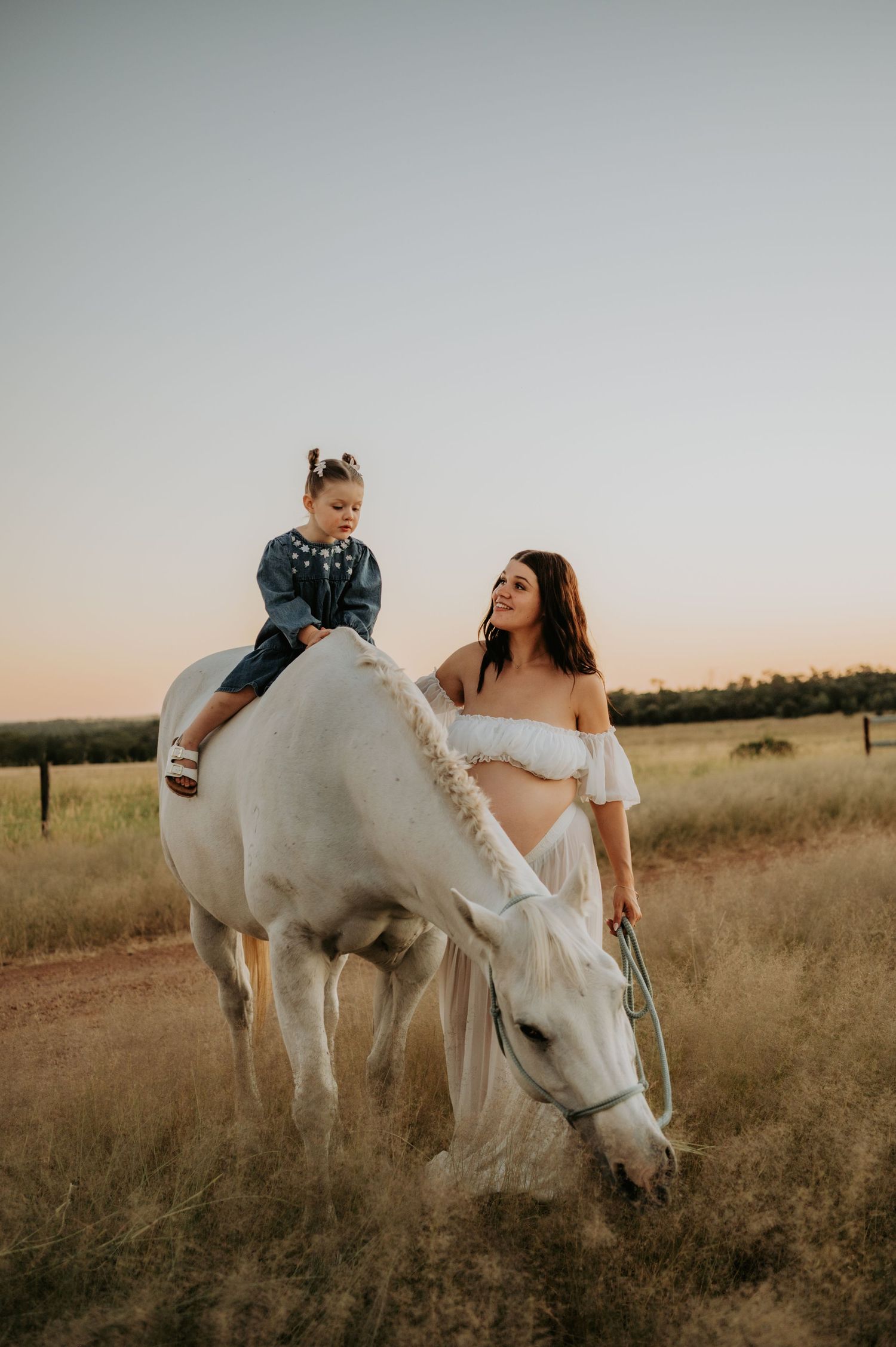 Pregnant woman in white dress leads a white horse with a young girl sitting on its back in a golden field at sunset.