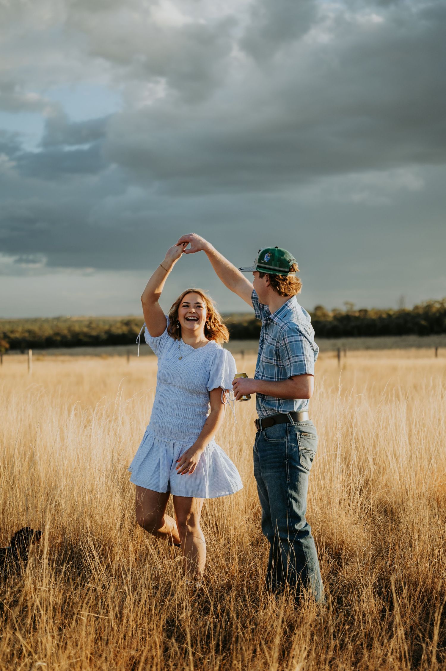 A joyful couple dancing in a golden wheat field under dramatic storm clouds in a rural countryside setting.
