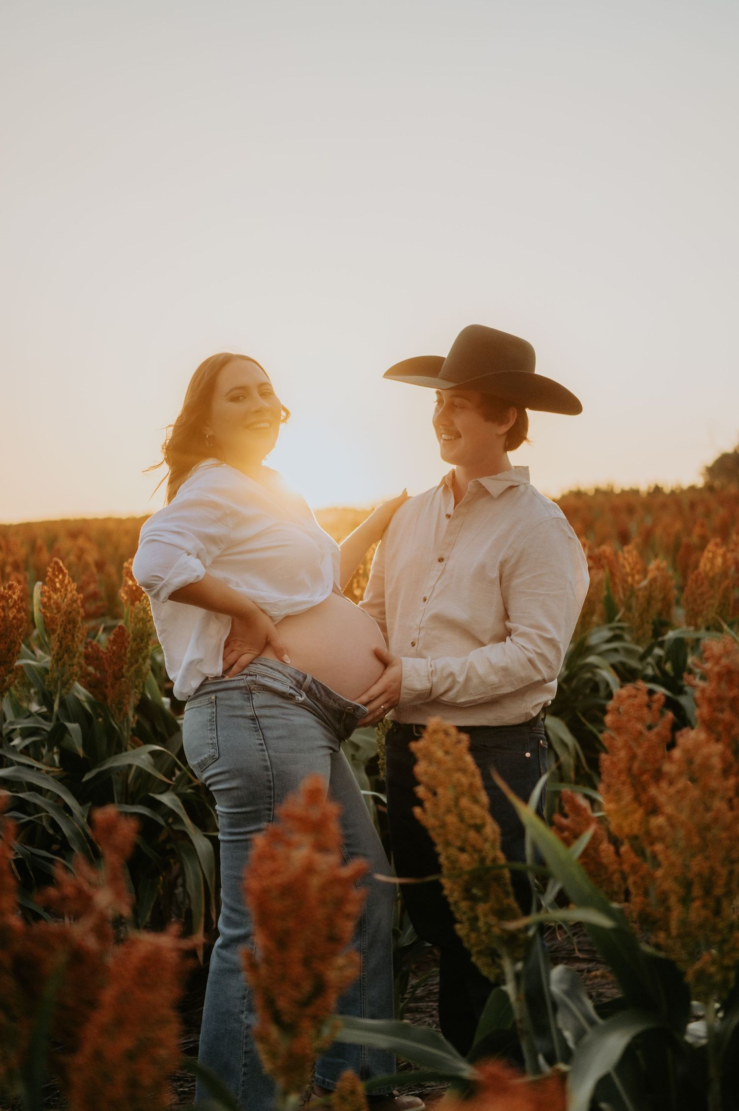 Pregnant couple poses in golden hour sunlit field, woman in white shirt and jeans, man in cowboy hat touching baby bump.
