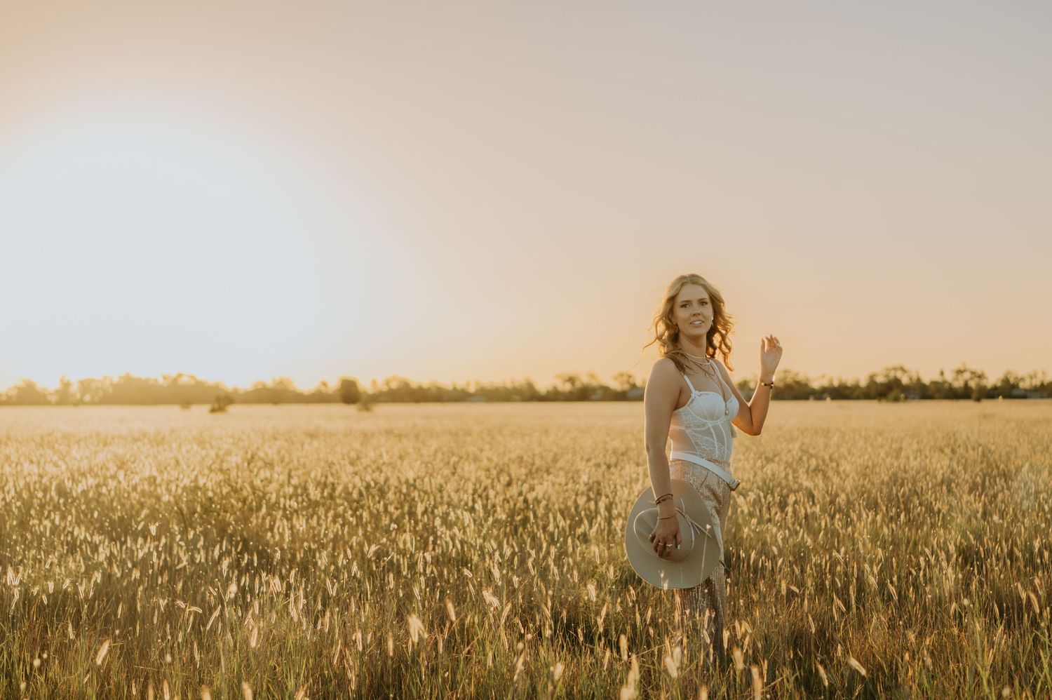 Woman in gray dress standing in golden wheat field at sunset, waving and smiling in warm sunlight.
