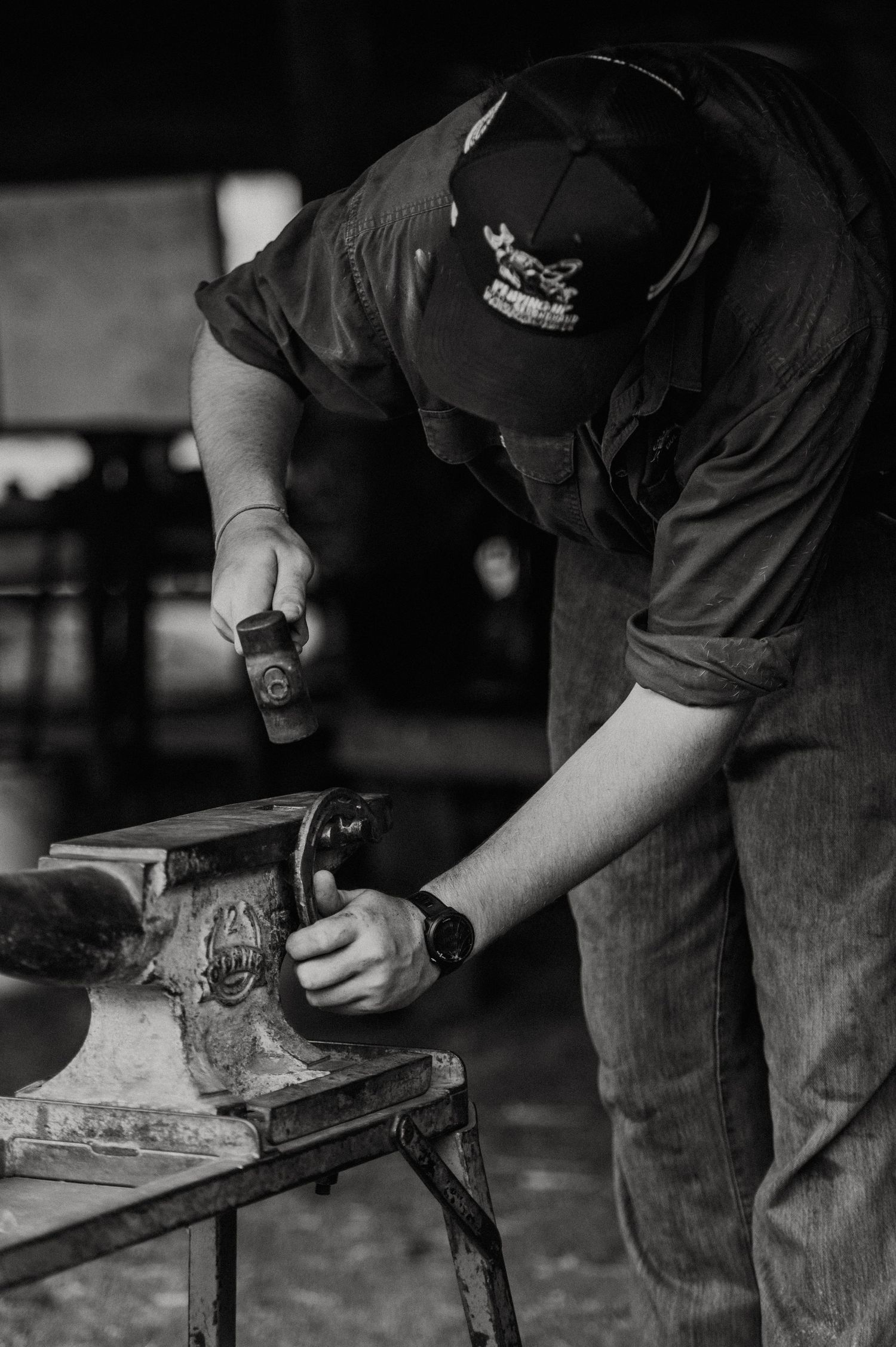 Black and white photo of a craftsman using a hammer at a workbench vise in a workshop.